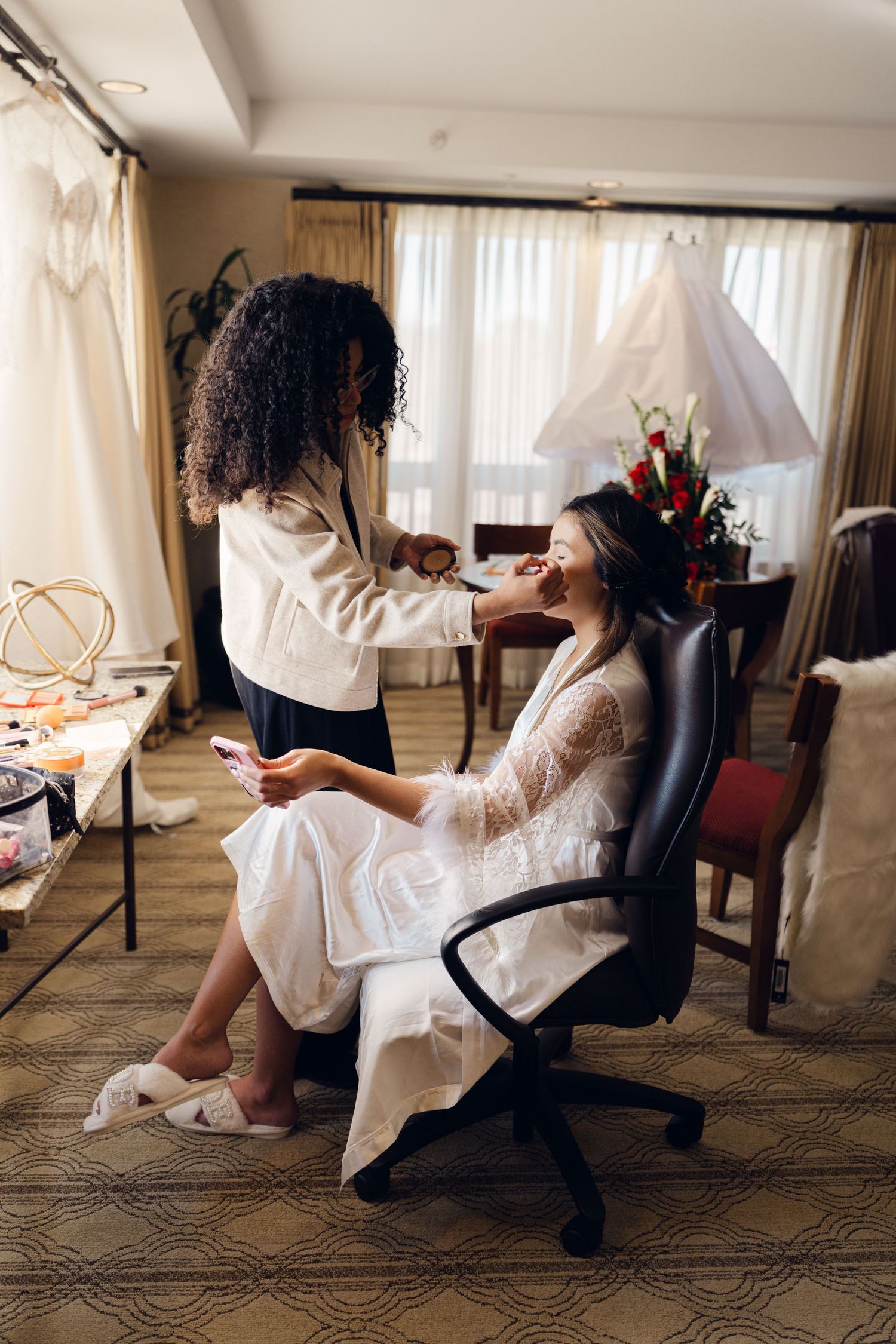 Makeup artist applying cosmetics to a smiling bride seated in a hotel room before her wedding.
