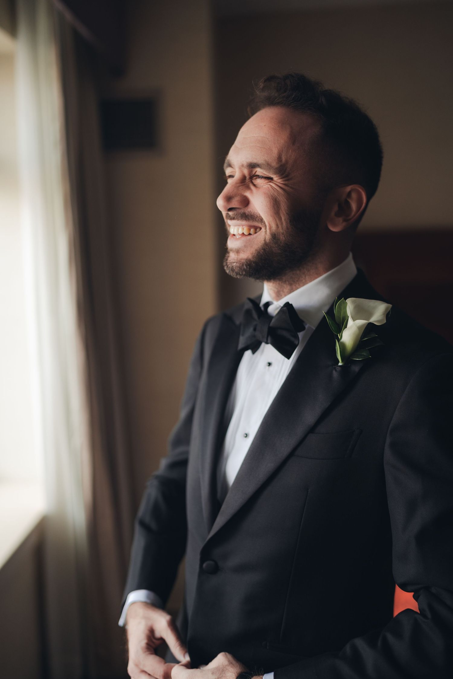 Handsome smiling groom in black tuxedo with bow tie and white calla lily boutonniere by window.