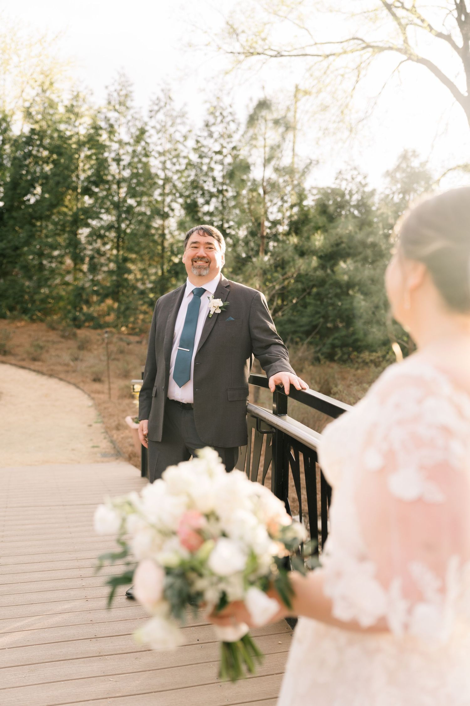 Groom smiling joyfully at bride during outdoor first look on a wooden bridge surrounded by trees.