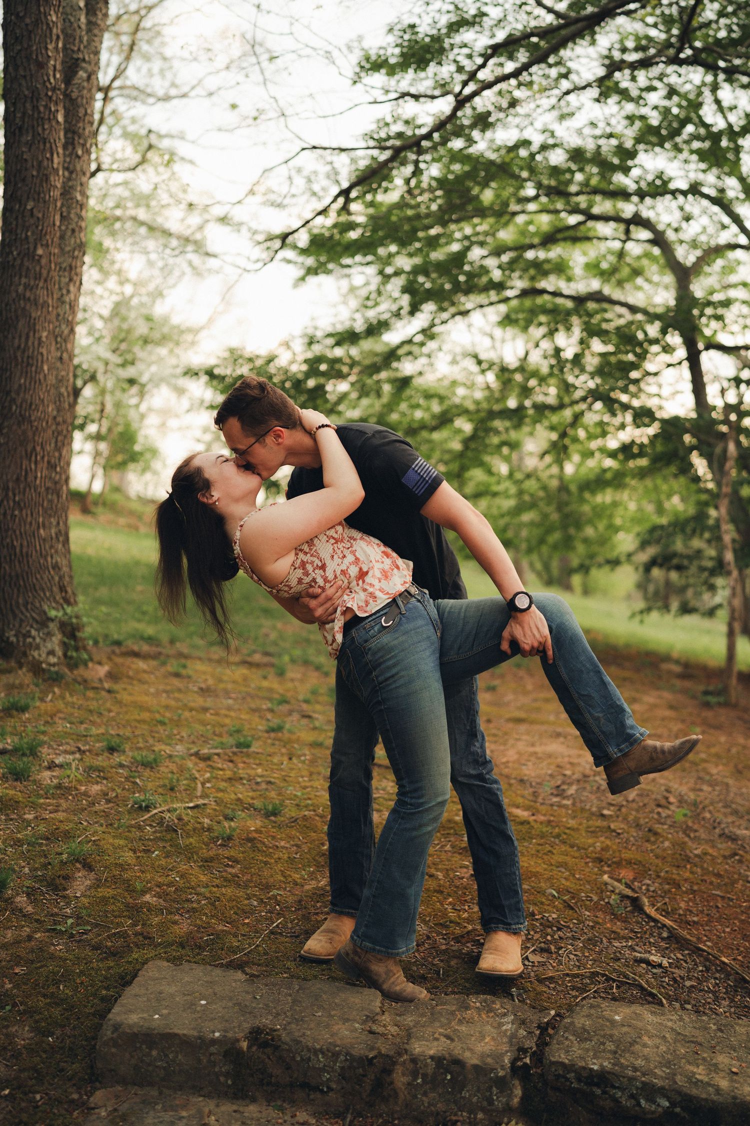 Man lifting woman for a kiss among trees on mossy rocks in a lush park.