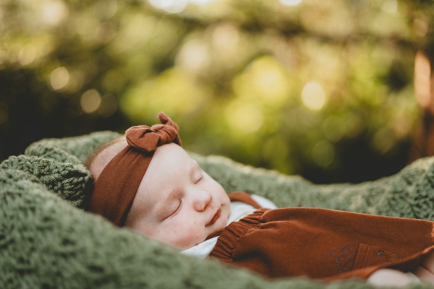 Peaceful sleeping newborn in brown bow headband rests on green blanket in soft bokeh light.