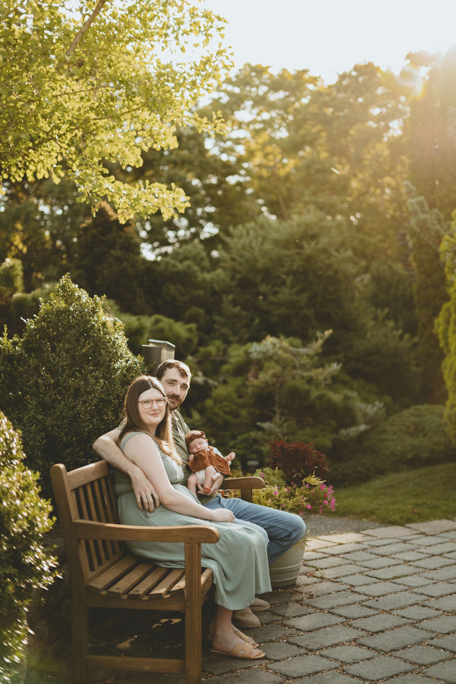 A couple sits together on a wooden garden bench holding their newborn in warm golden sunset light.