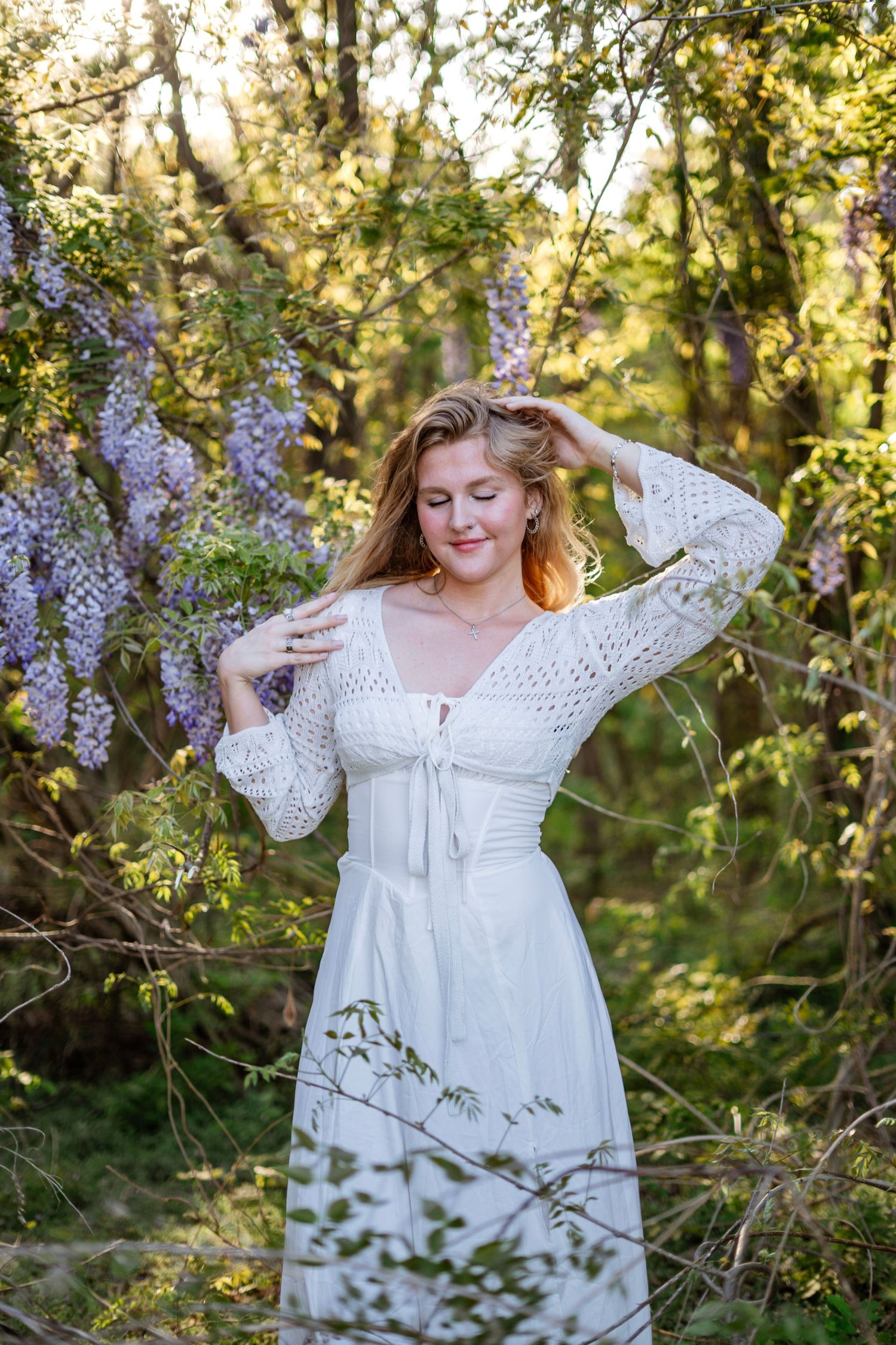 A blonde woman in a white lace dress poses with hands in hair near purple wisteria blooms.