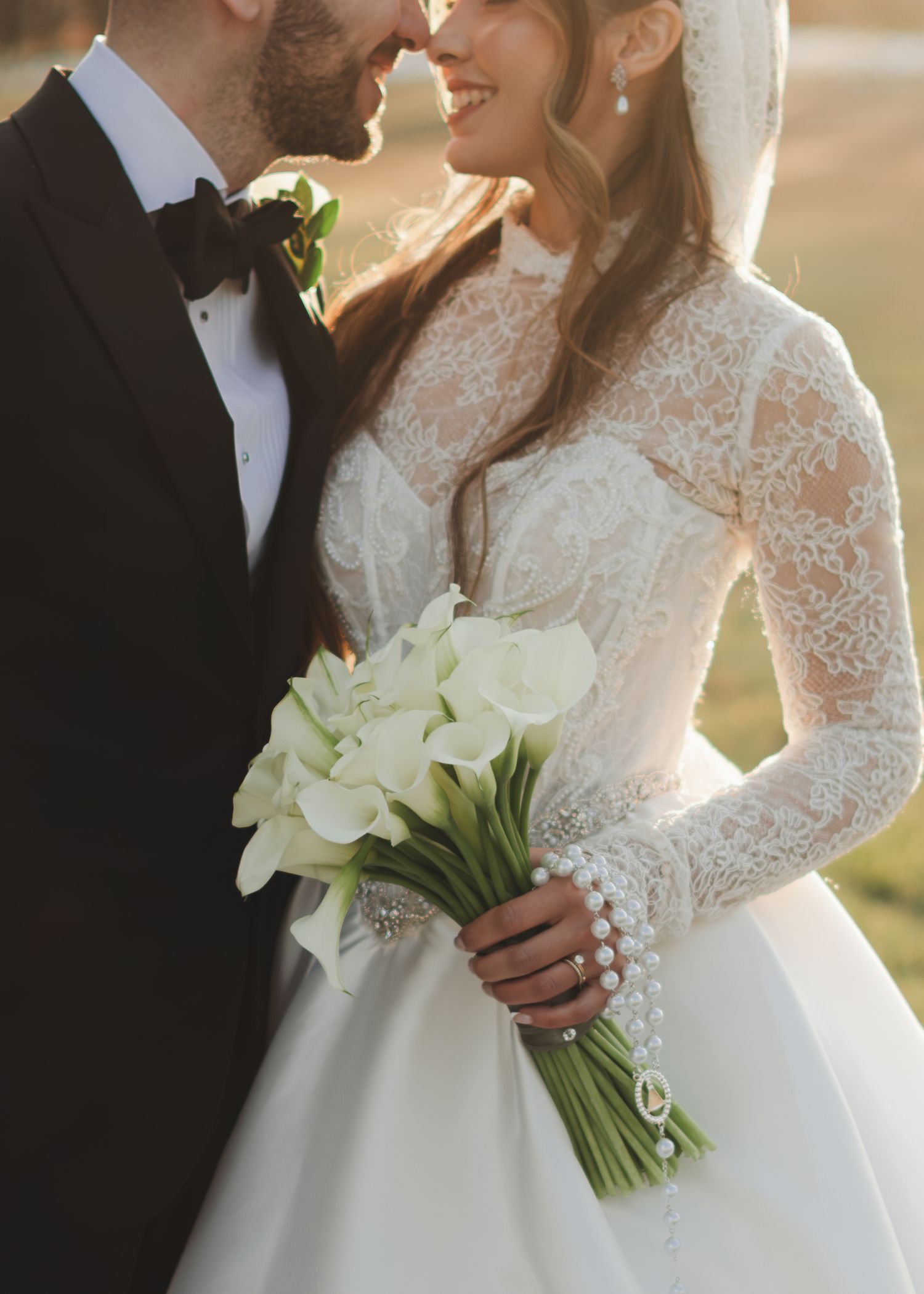 Bride in long-sleeve lace wedding gown holding elegant white calla lily bouquet close-up.