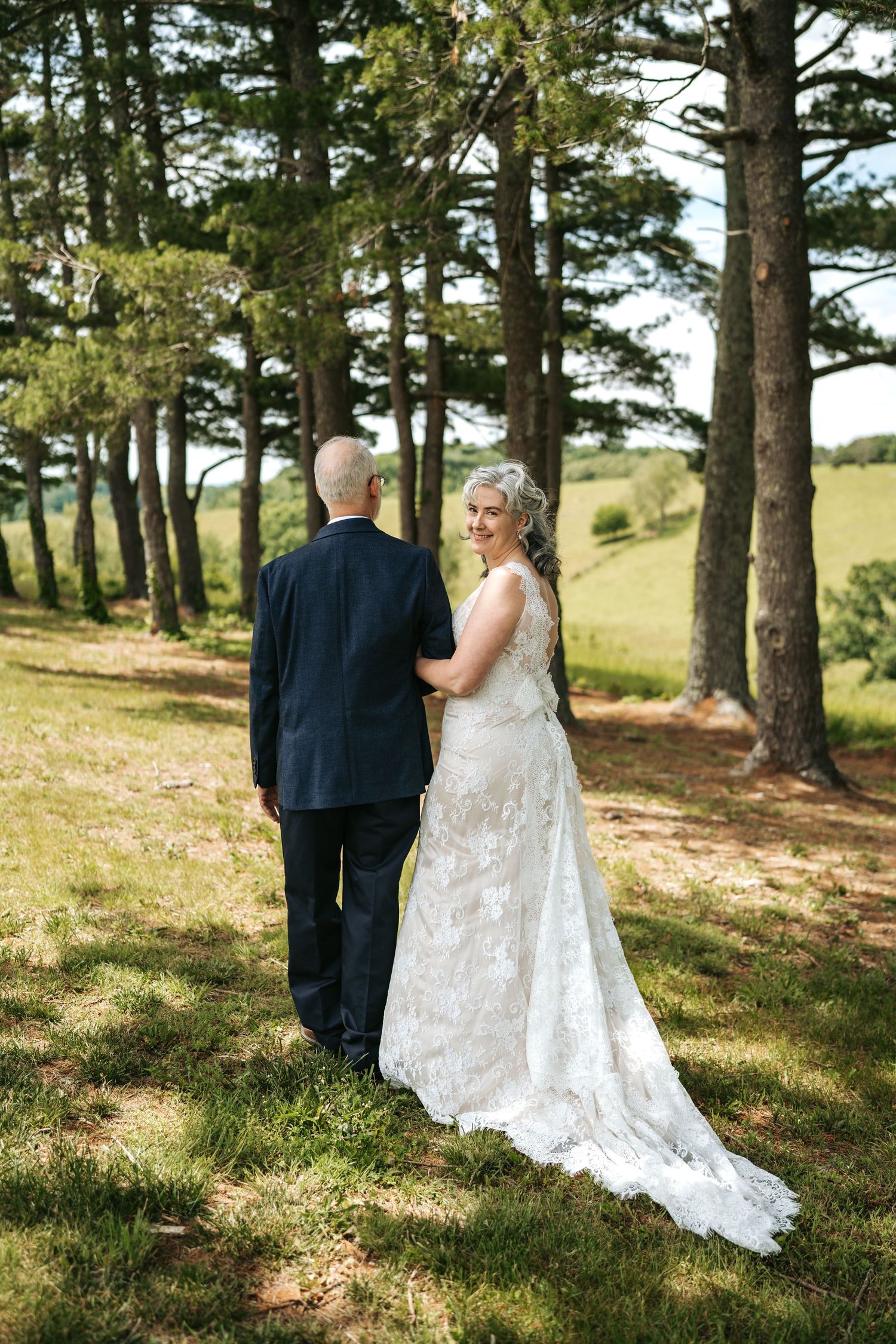 Bride in lace gown smiles over shoulder while walking with groom through sunlit pine forest on wedding day.