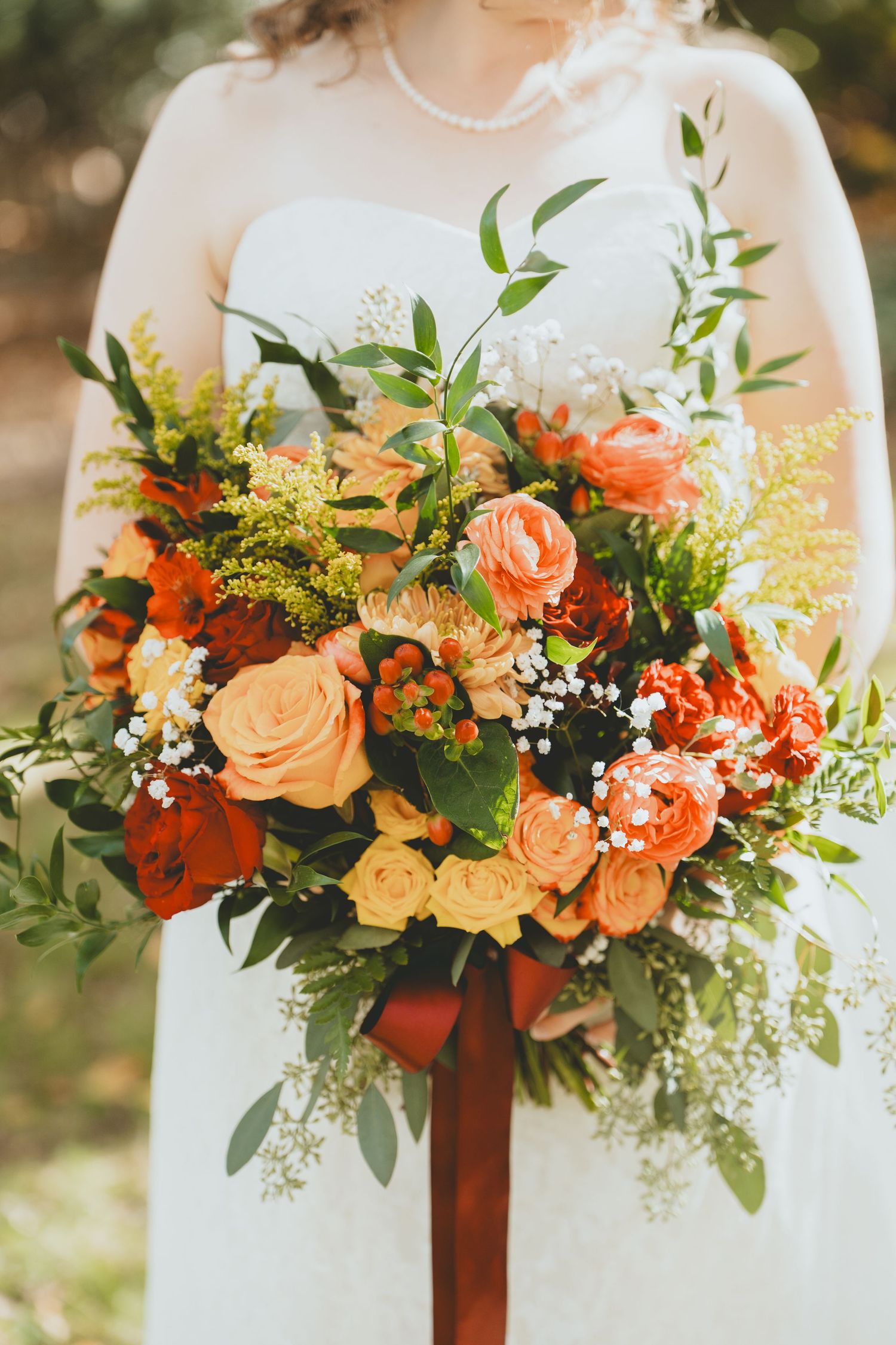 Bride holding a lush fall wedding bouquet with orange, red, and yellow roses with greenery.