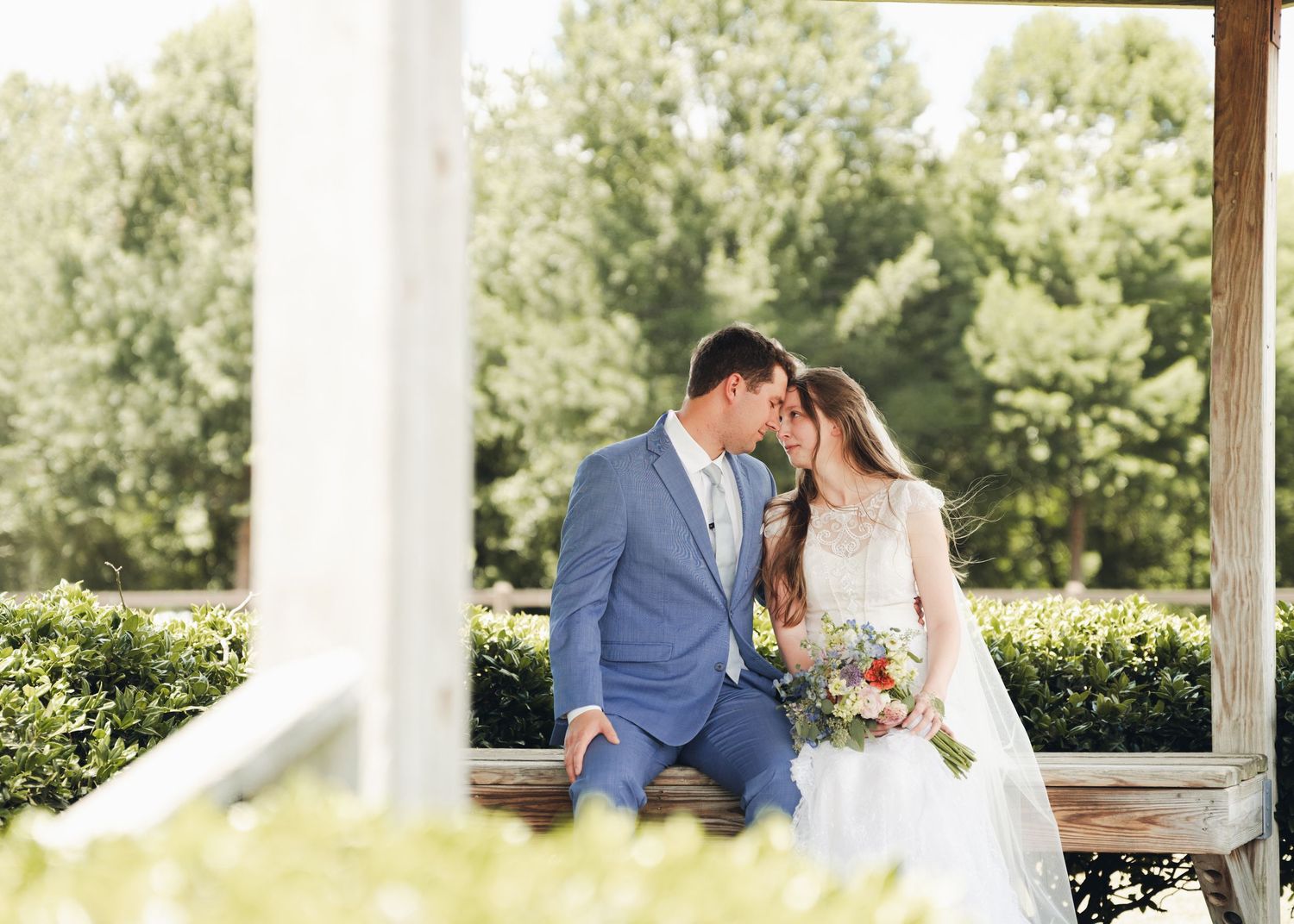 Bride and groom share a romantic forehead touch while seated outdoors, surrounded by lush greenery on their wedding day.