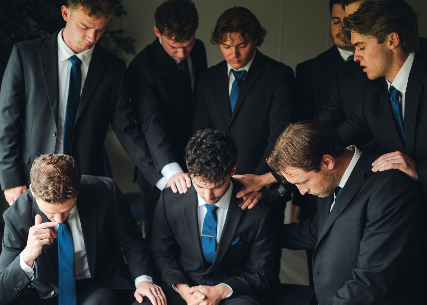 Group of young men in dark suits with blue ties gathered closely together in a formal prayer circle.
