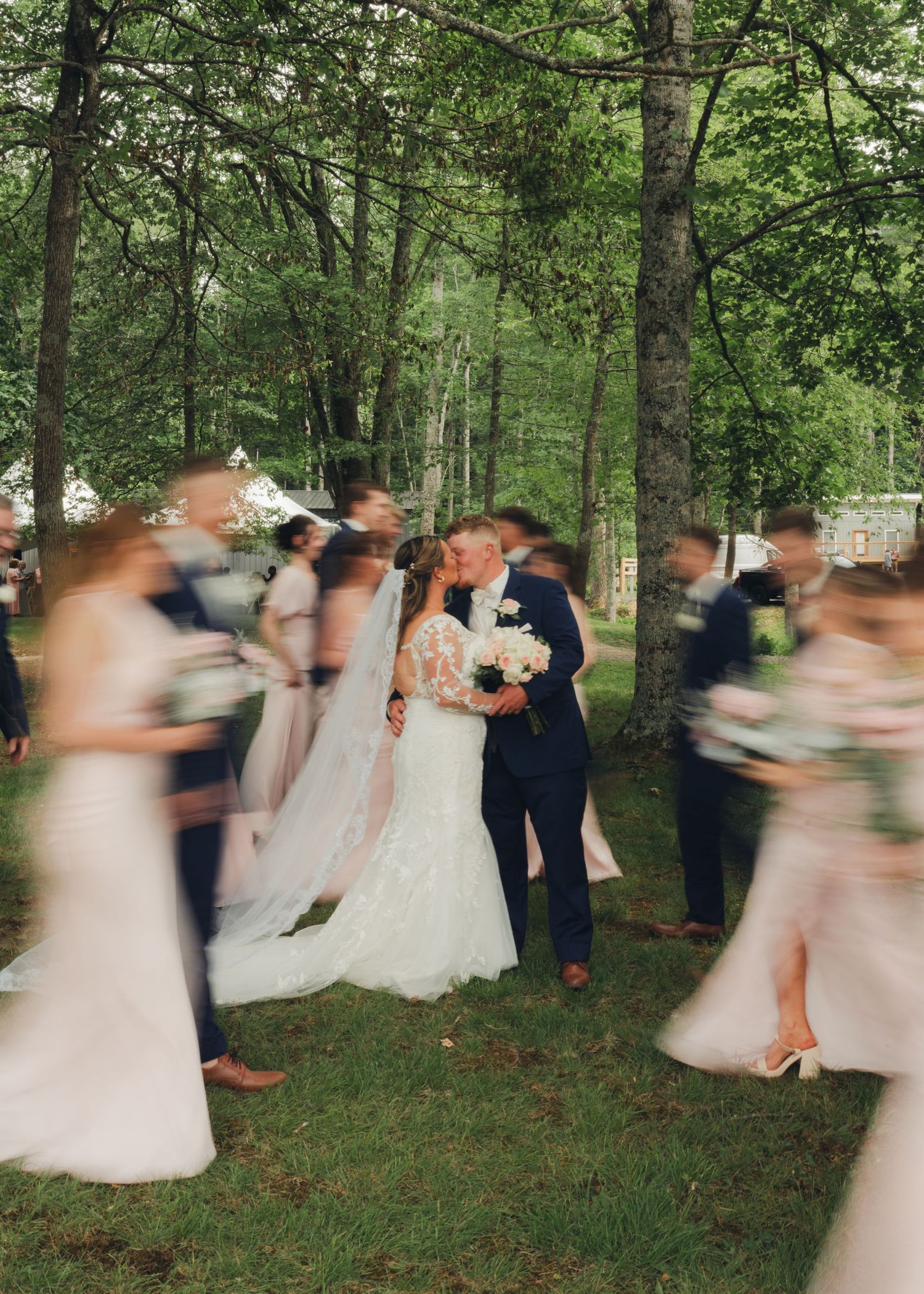 Bride and groom share a kiss surrounded by blurred wedding party guests in a lush green wooded outdoor setting.