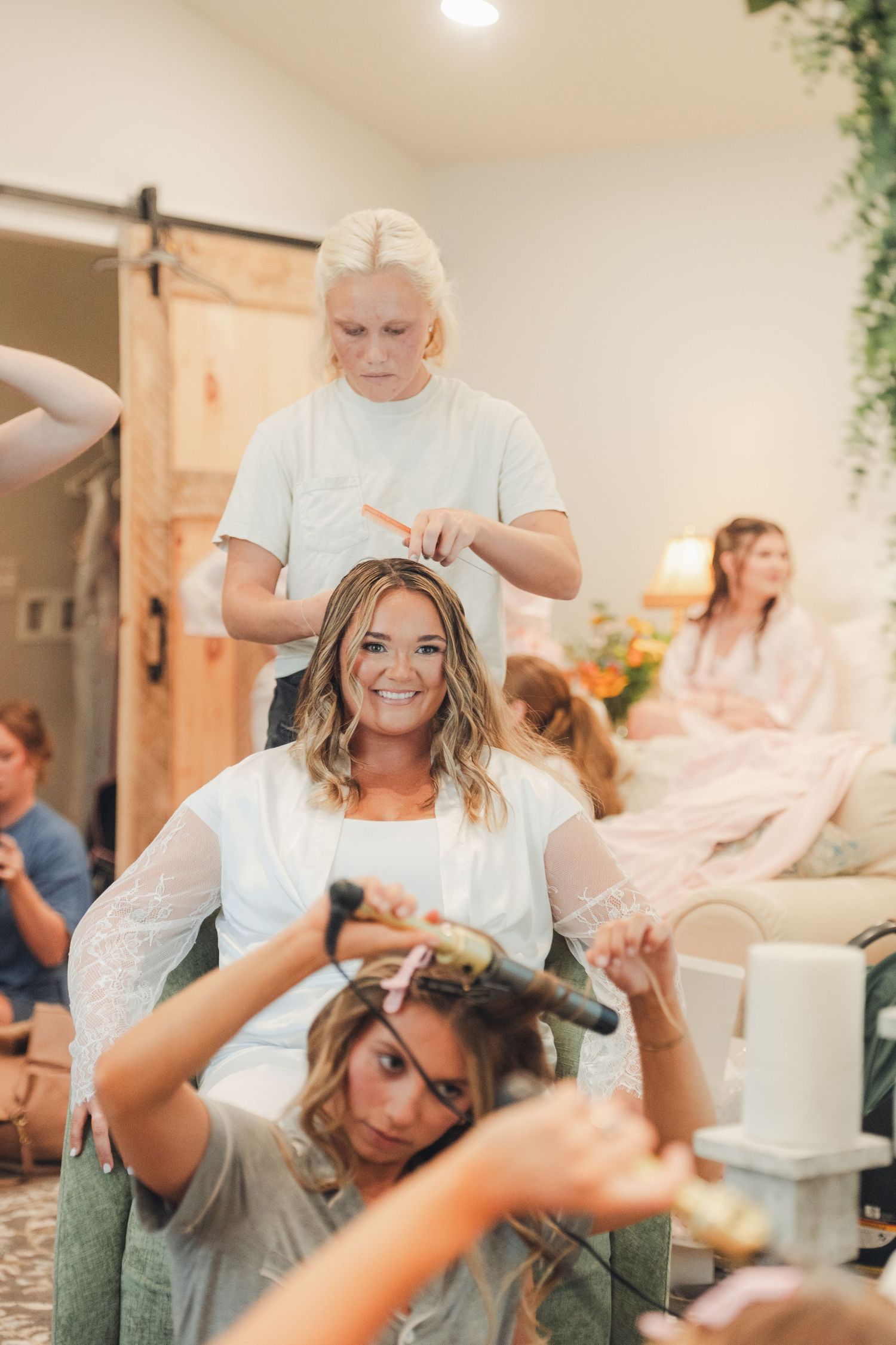 Bride smiling while getting hair styled by two stylists in a bright bridal suite on wedding day.
