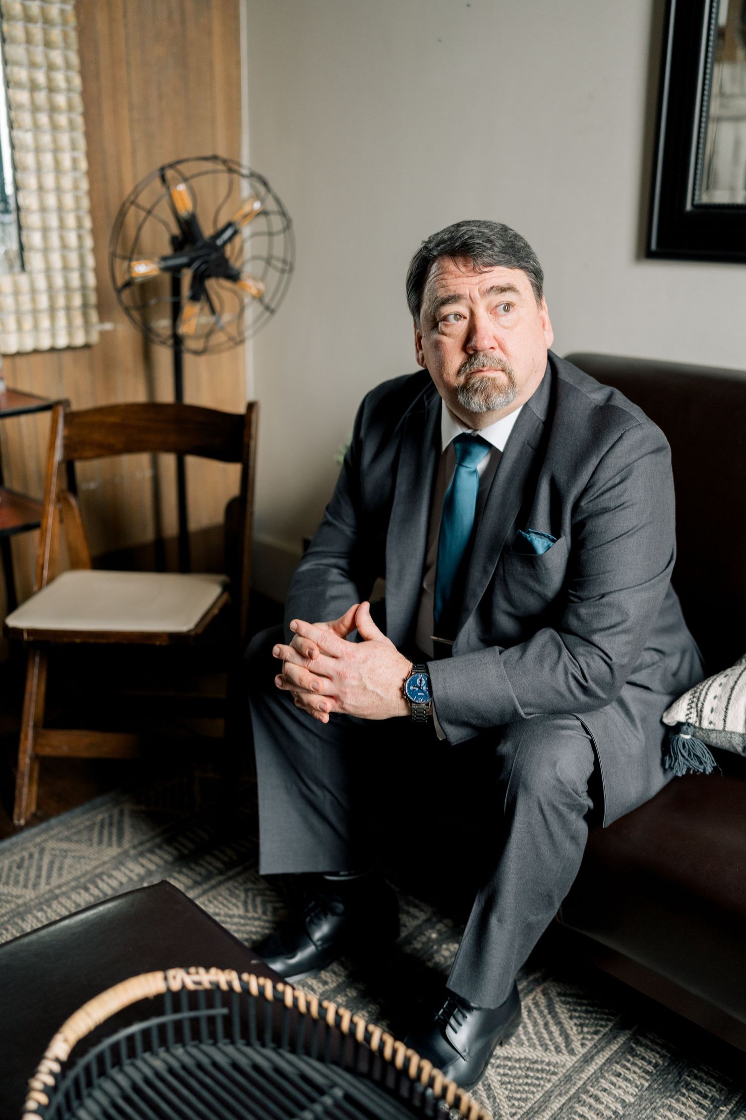 Older gentleman in gray suit with teal tie sits on leather sofa in vintage-styled room.