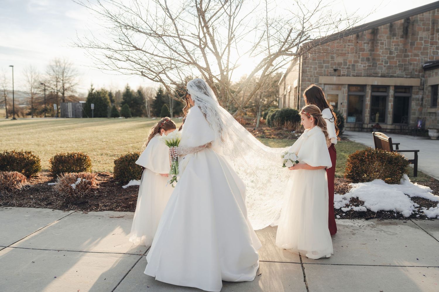 Bride in white ball gown with long veil assisted by flower girls and bridesmaid outside church.