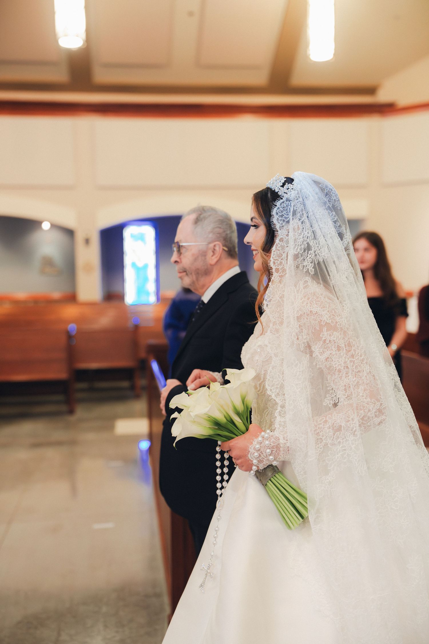 Bride in lace wedding gown holds white calla lily bouquet while walking down church aisle with father.