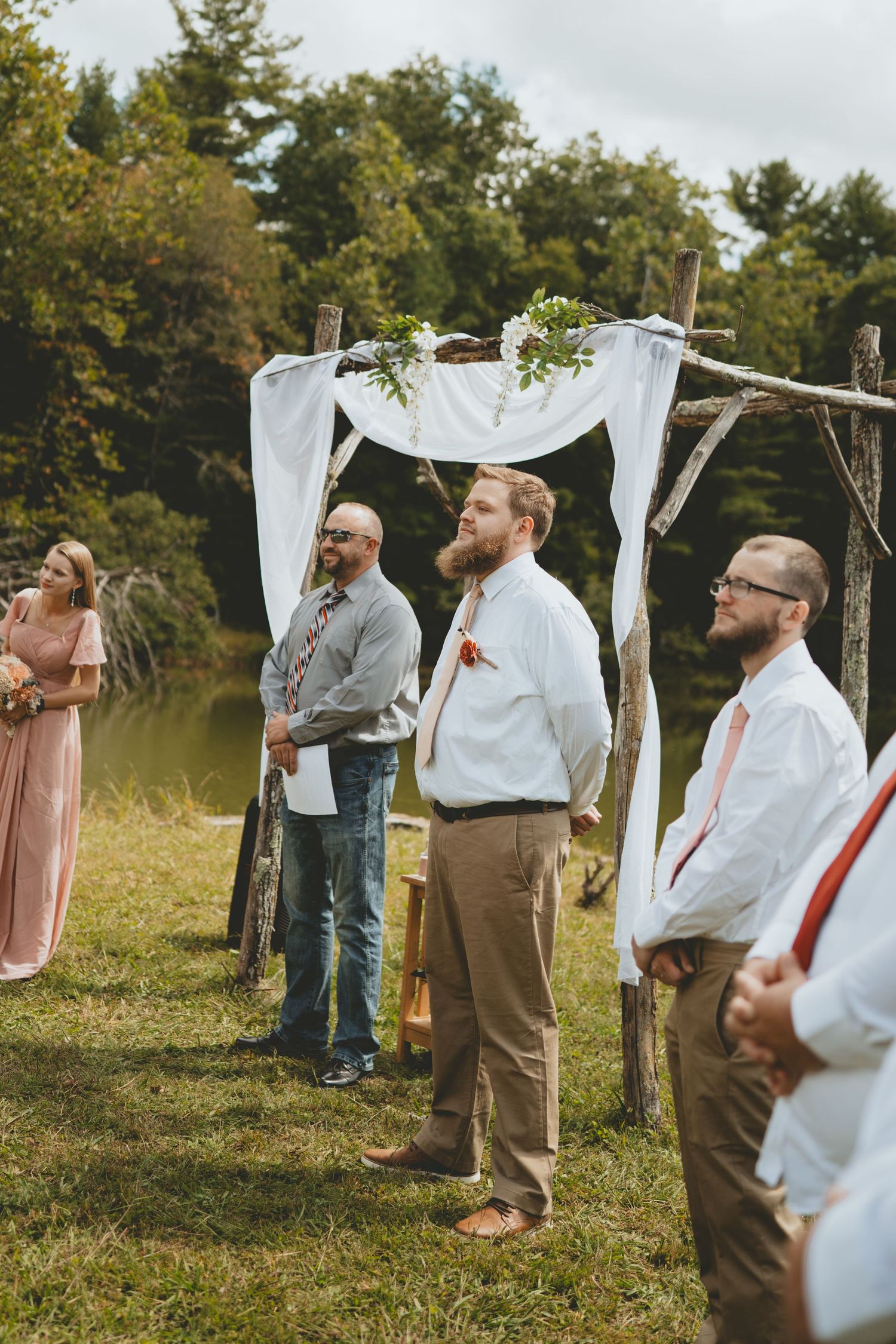 Groomsmen standing at attention near a rustic wooden wedding arch with white fabric by a pond.