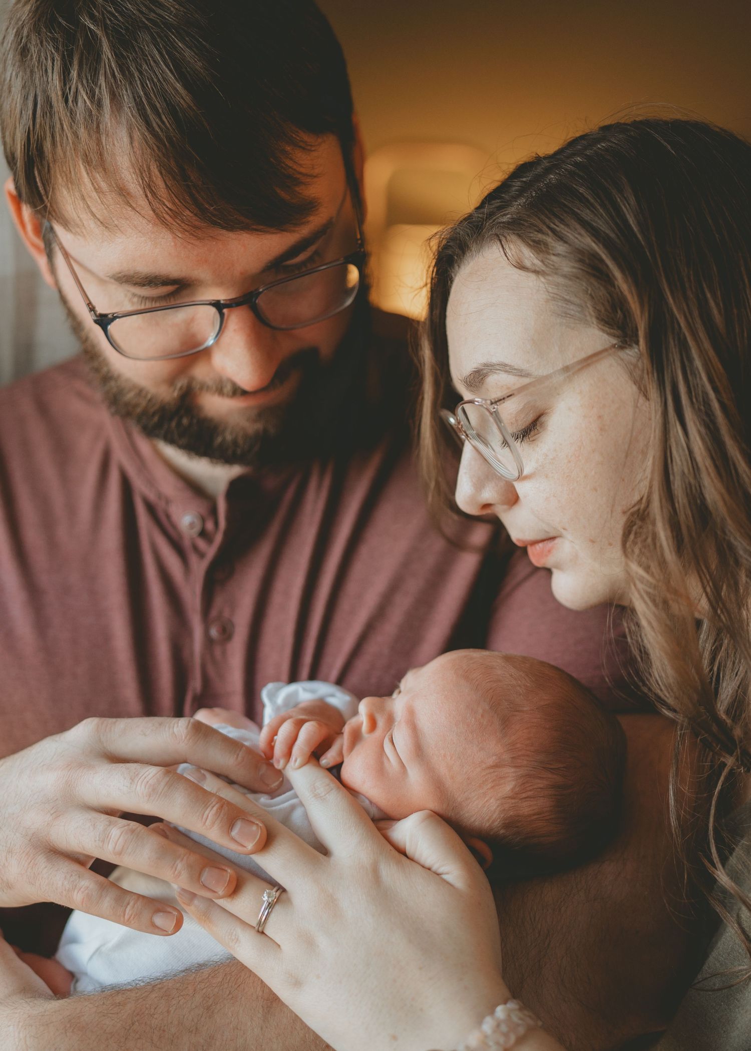 New parents with glasses gently cradle their newborn baby together in a warm, intimate home setting.