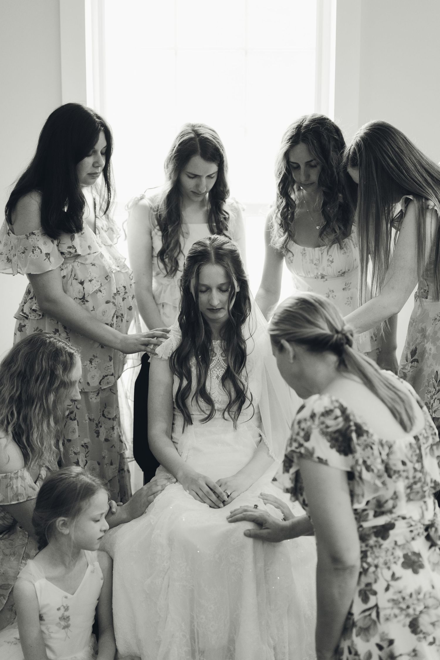 Black and white photo of bridesmaids helping bride with veil in bright window light.