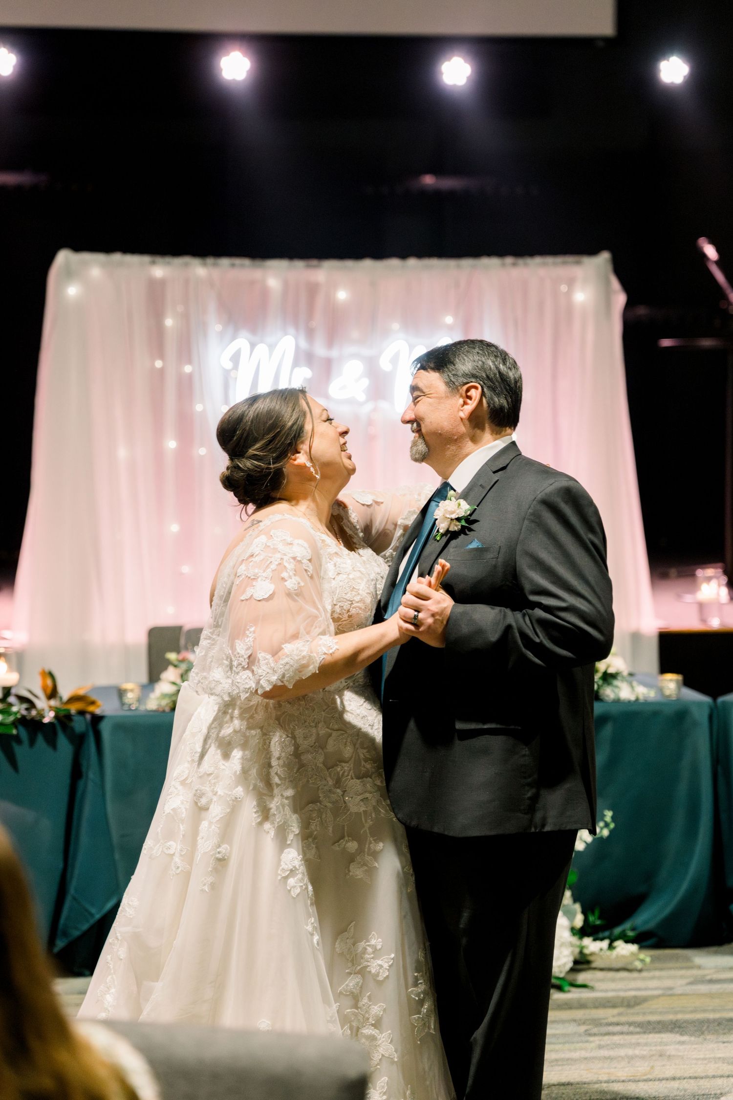 Bride and groom share joyful first dance at elegant wedding reception with monogram backdrop and stage lighting.