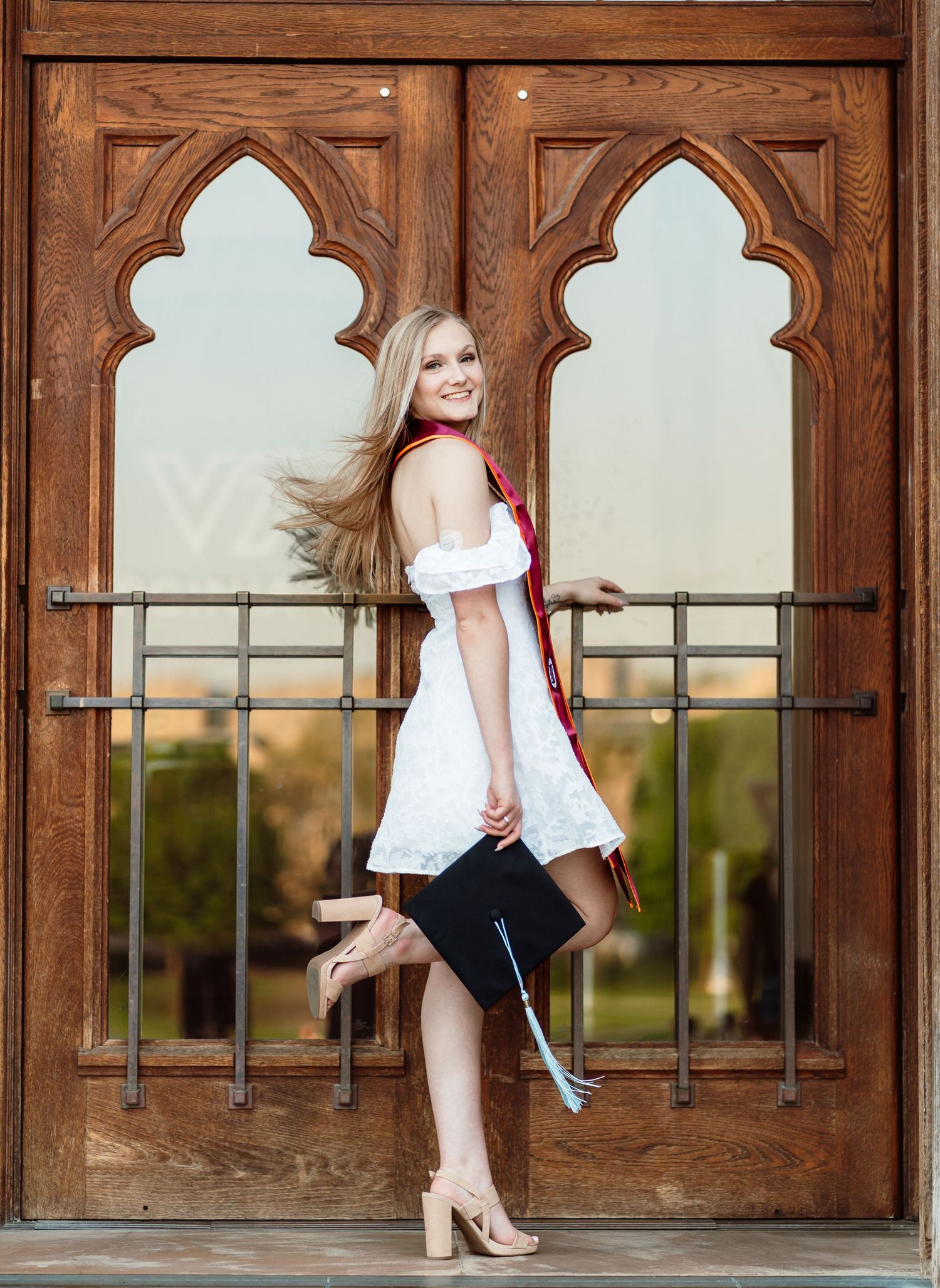 Smiling blonde graduate in white dress holding cap beside ornate wooden doors at golden hour.