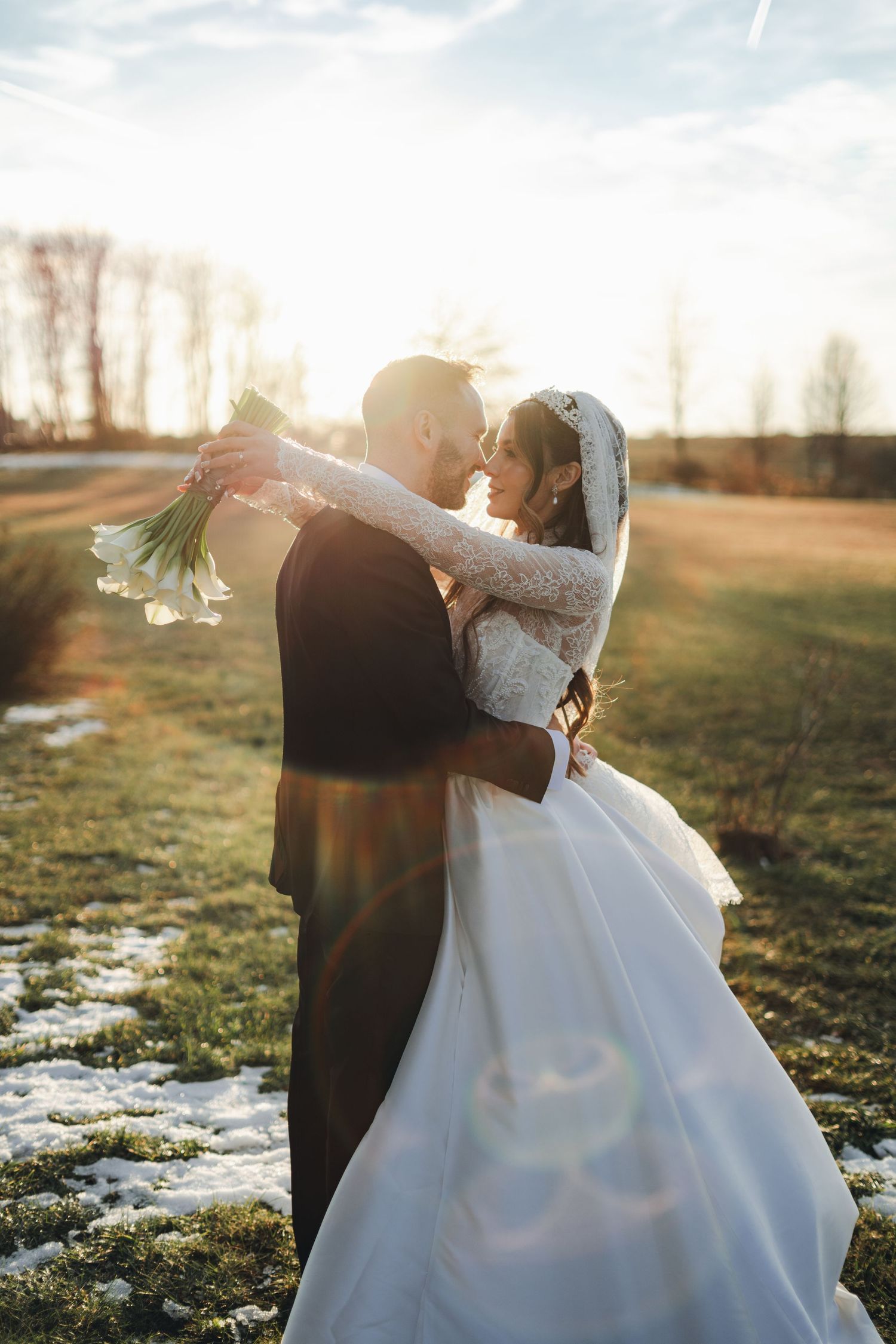 Bride and groom embrace at golden sunset in snowy field, bride holding white calla lily bouquet.