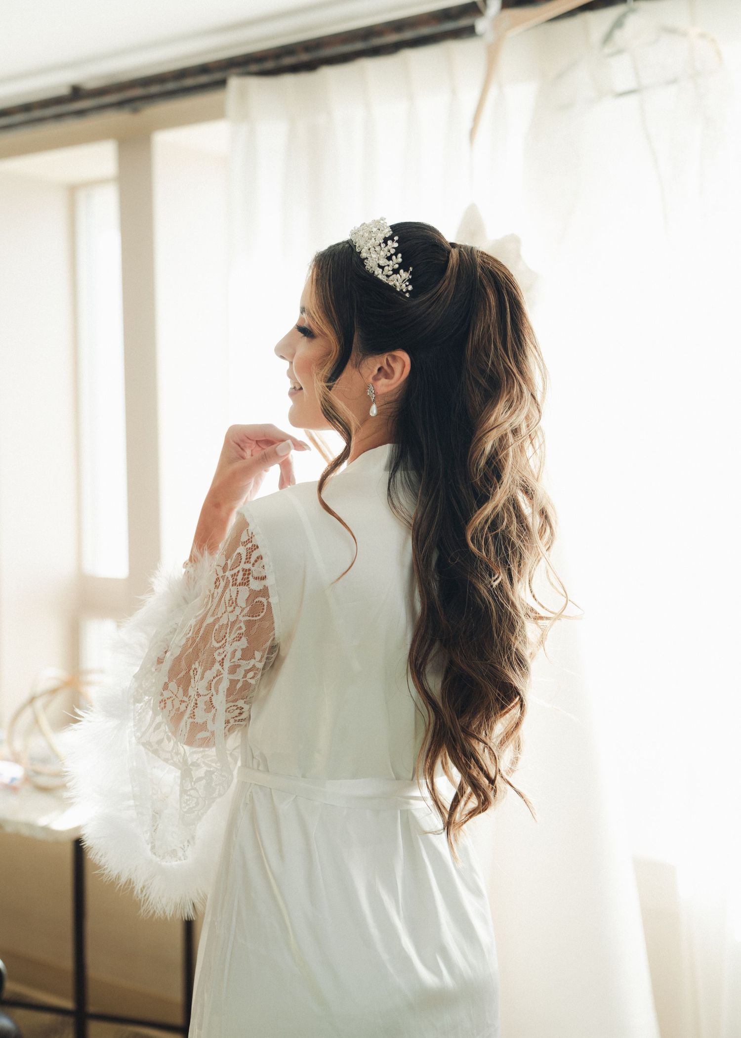 A bride in a white lace robe with a pearl hair accessory admires her long wavy hair by a window.