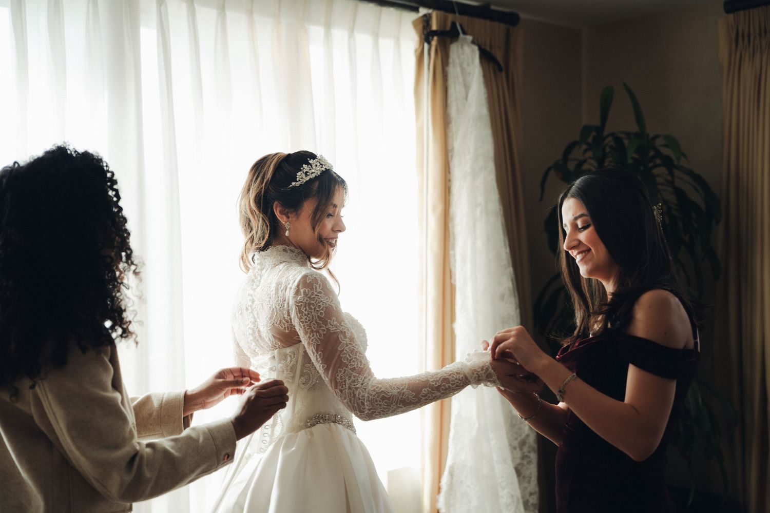 Bride in lace gown and tiara smiling as two women help her prepare near a bright window.