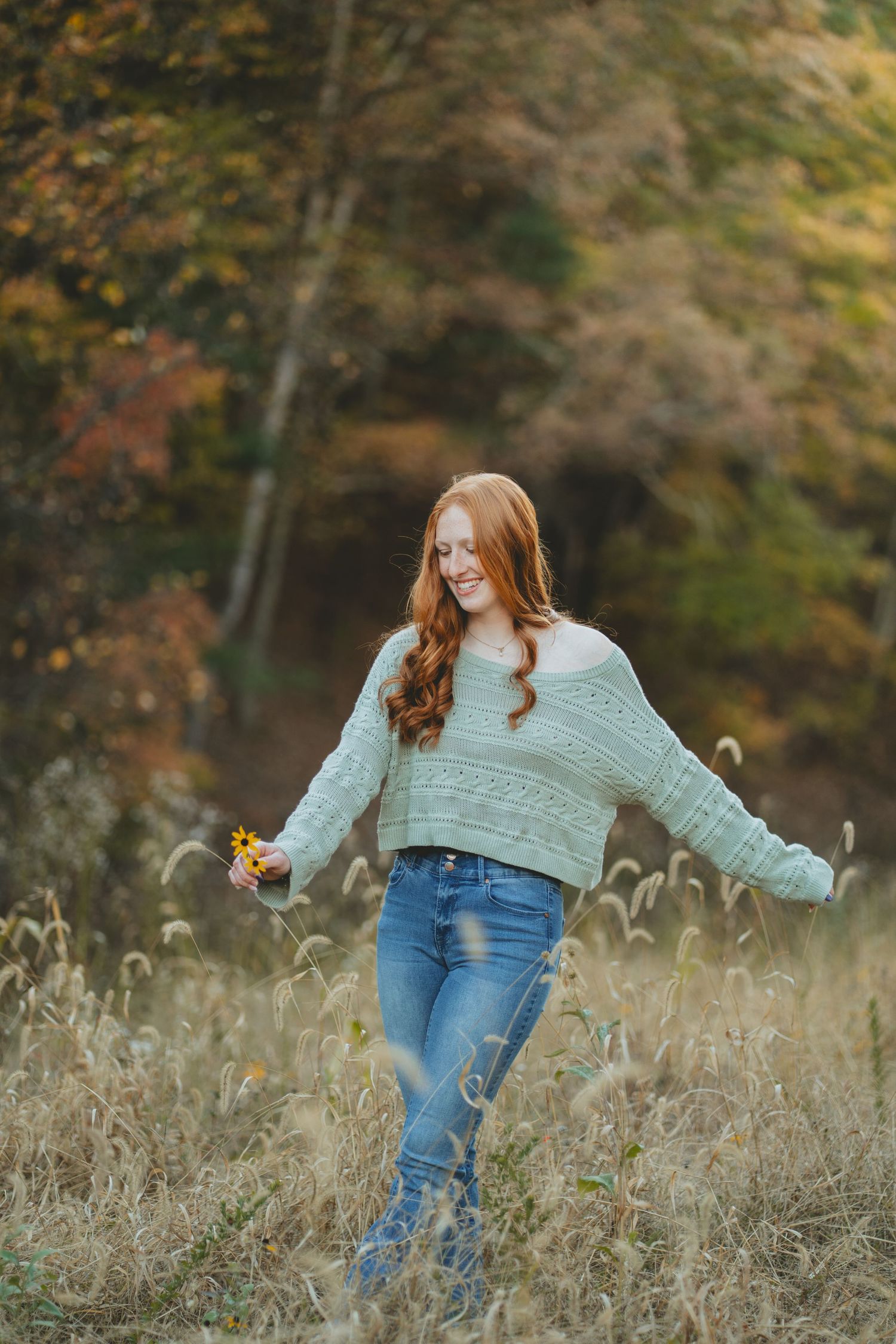 Smiling redhead girl in mint sweater and jeans walking through tall autumn grass.