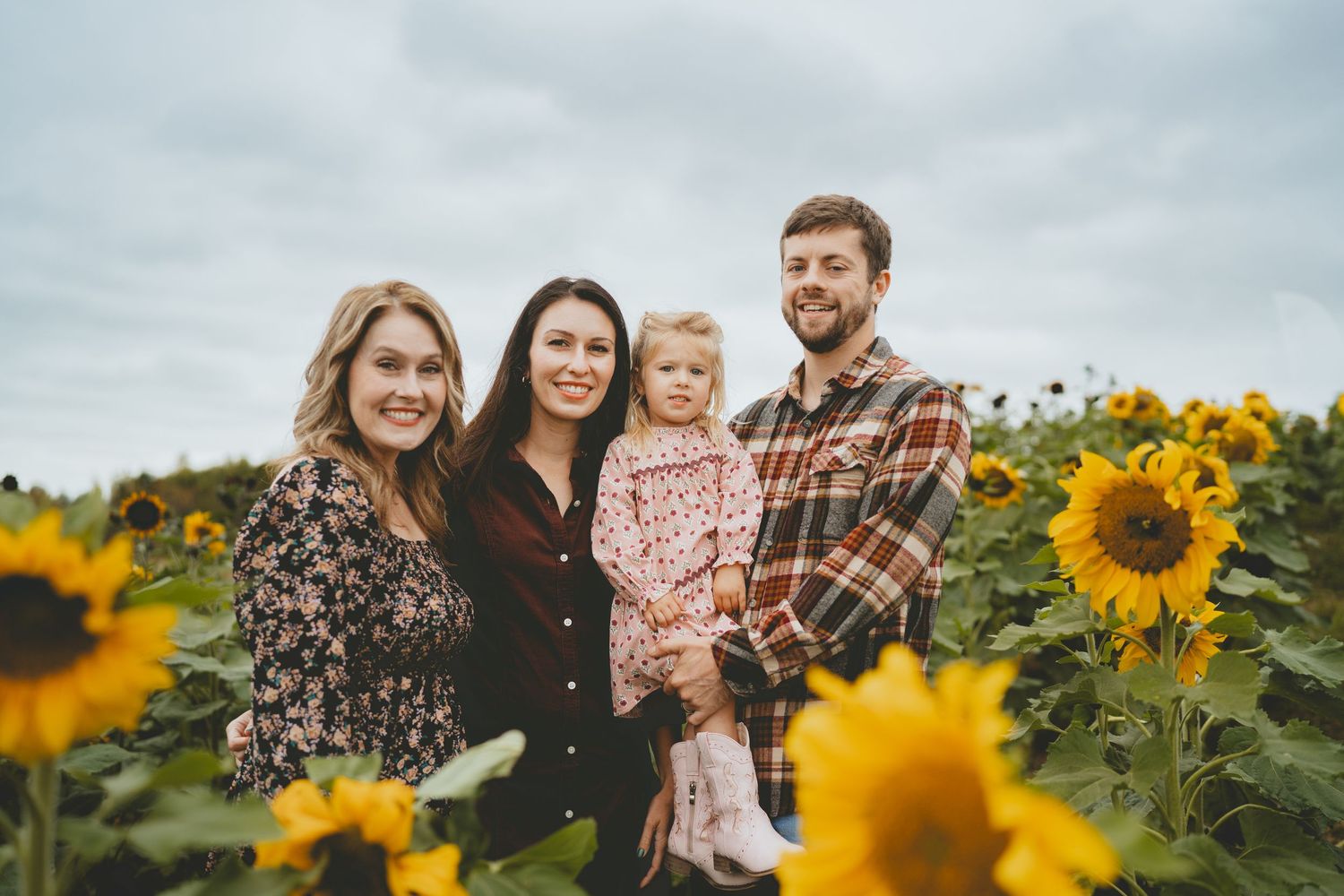 Family of five smiling in a vibrant sunflower field on a cloudy day.