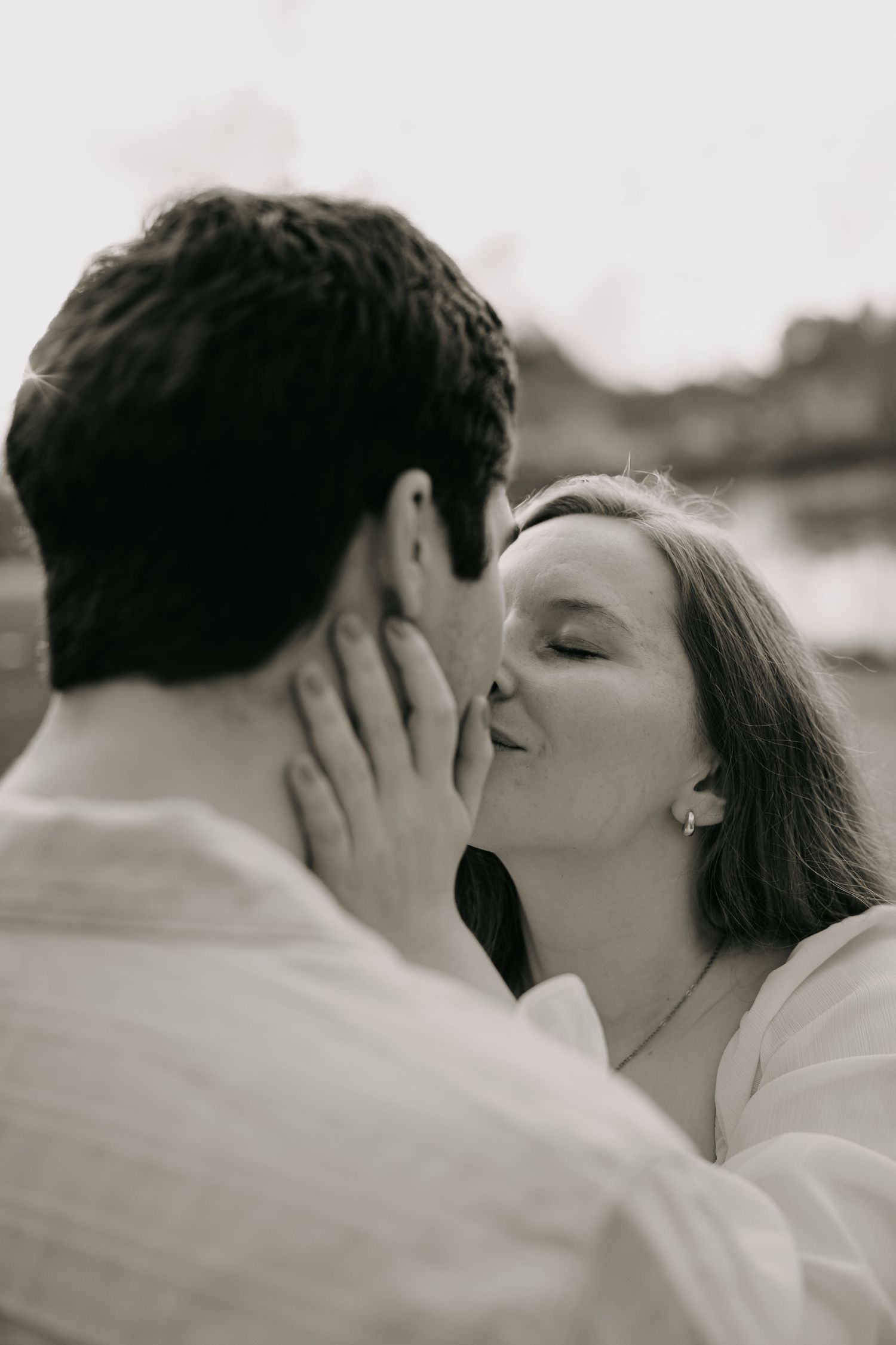 Black and white romantic photo of couple sharing a kiss outdoors near a serene waterway.