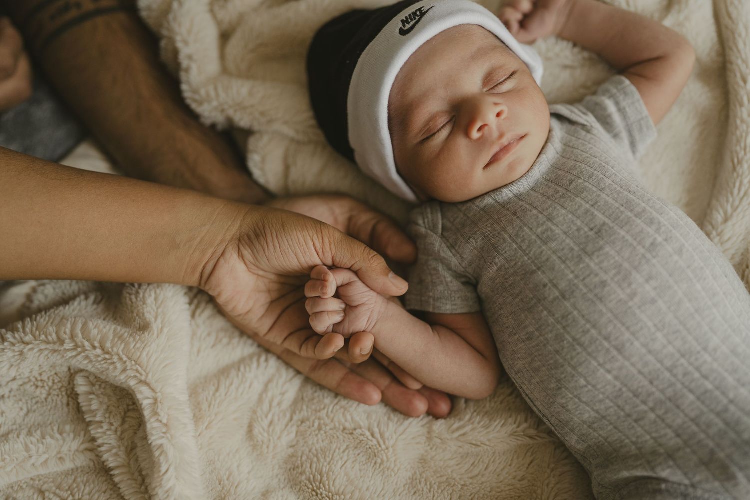 Peaceful sleeping newborn in gray swaddle and white hat holding parent's finger on cream blanket.