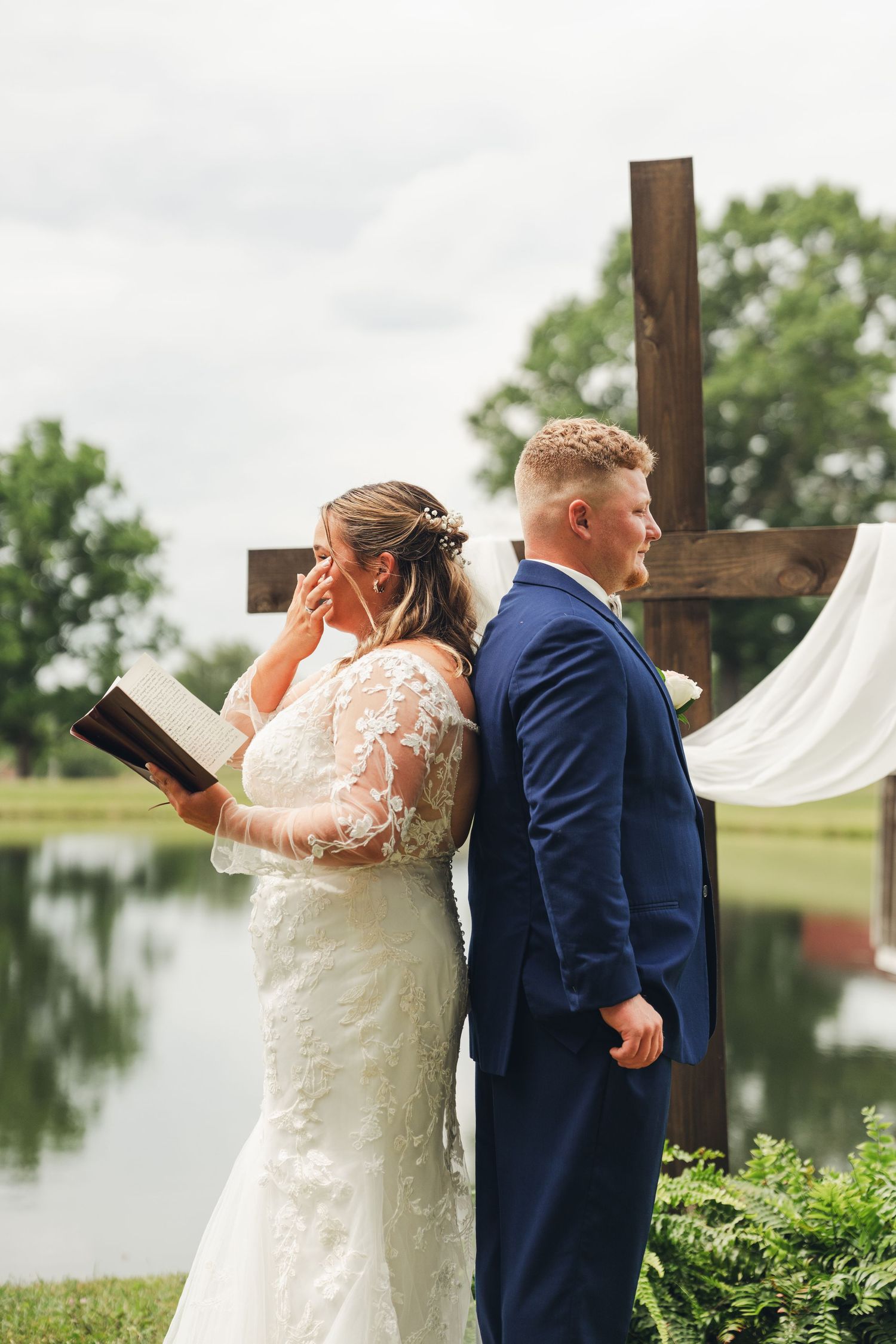 Bride and groom stand back-to-back during outdoor wedding ceremony near a wooden cross and pond.