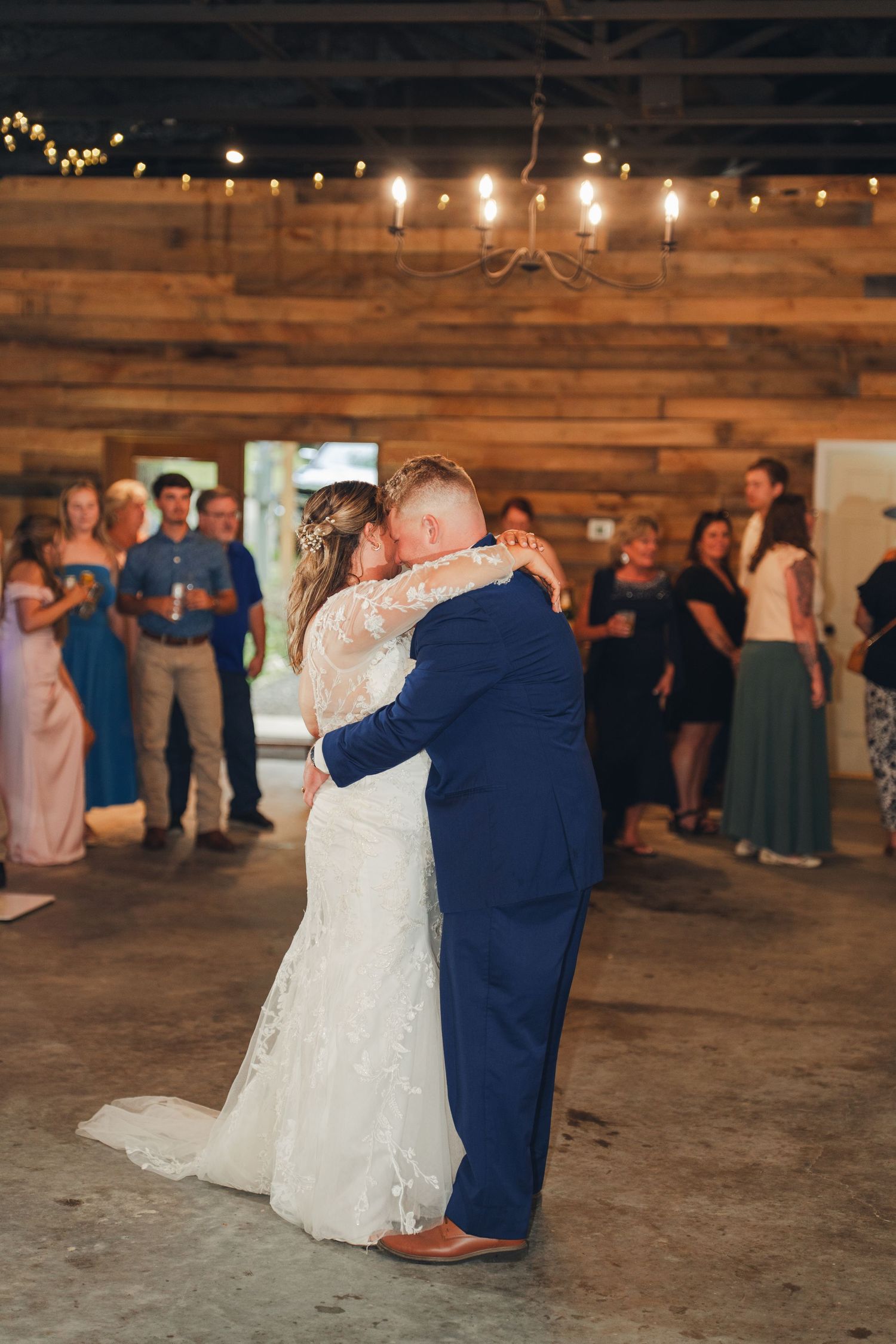 Bride and groom share first dance in rustic barn venue with string lights and chandelier, guests watching in background.