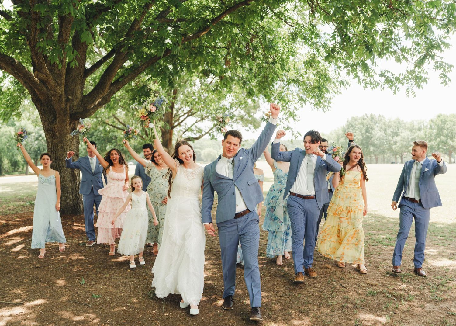 Joyful wedding party celebrating outdoors under a large tree, bride in white gown and groom in blue suit leading the group.