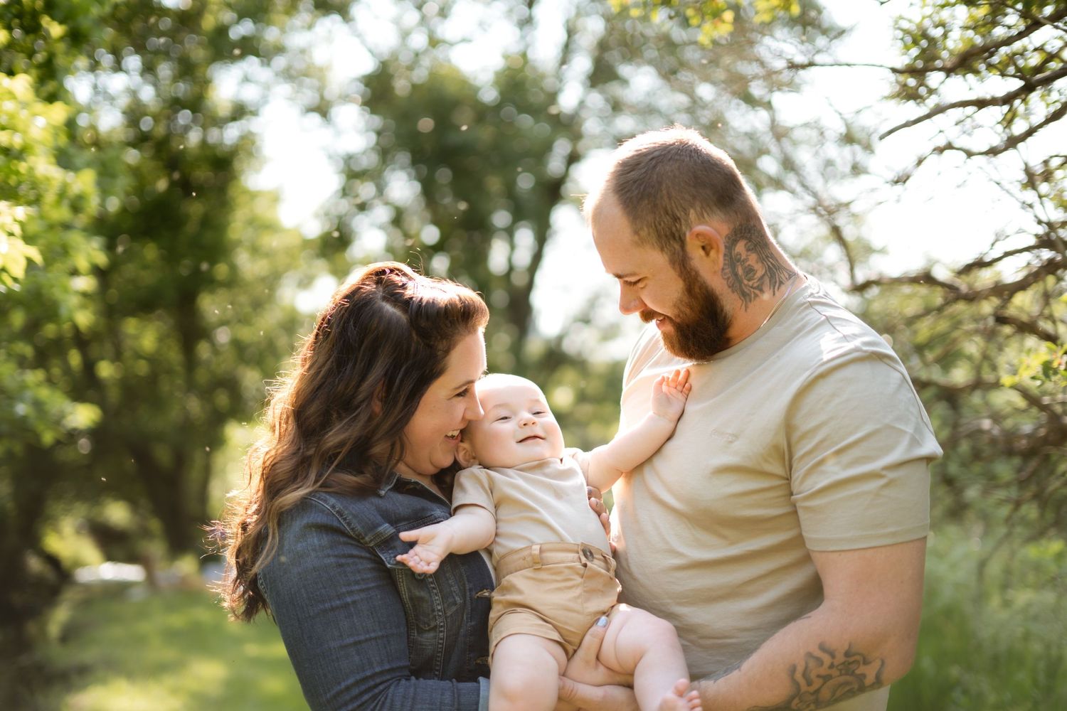 Happy young family laughing together while holding their baby in a sunlit outdoor setting.