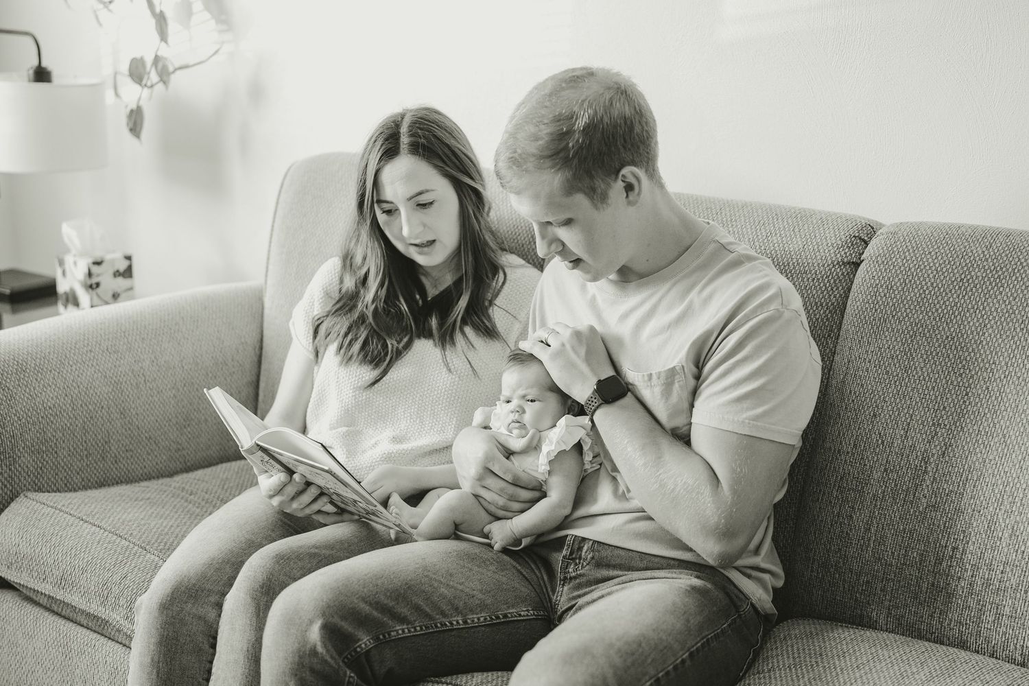 Black and white photo of parents sitting on couch reading to their newborn baby.