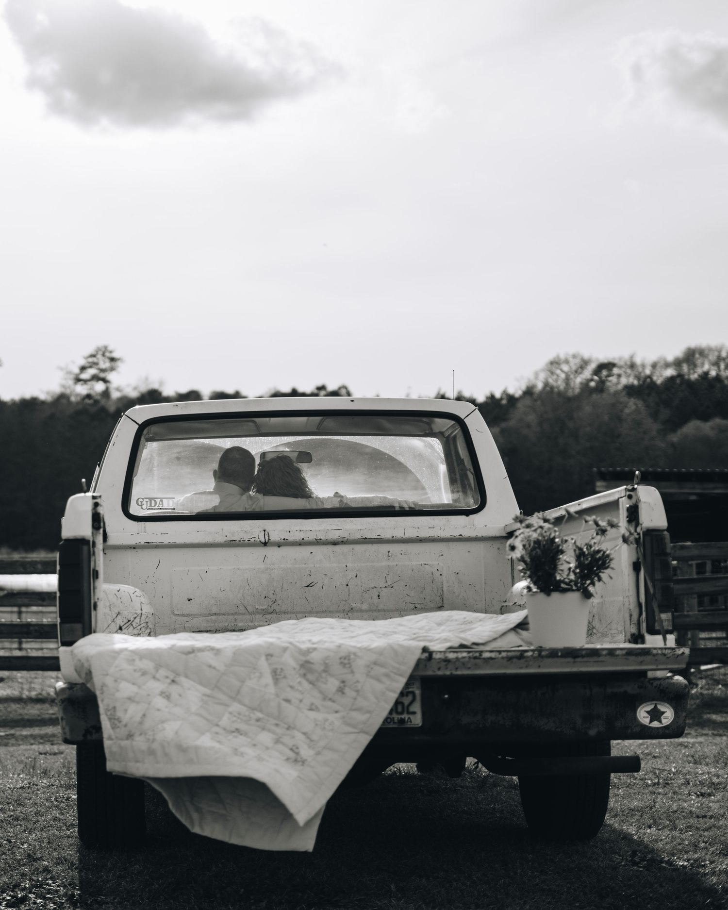 Black and white photo of a couple sitting inside an old pickup truck with a white blanket draped over the tailgate.