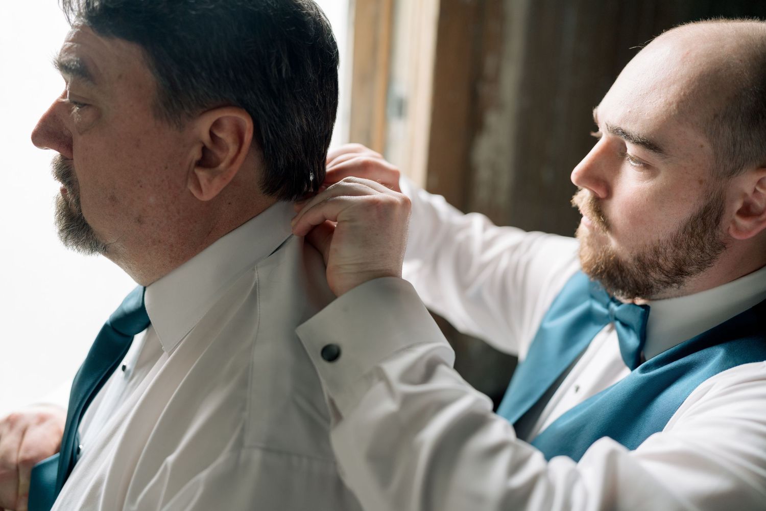 Groomsman helping groom with collar button wearing teal bow ties and white dress shirts.