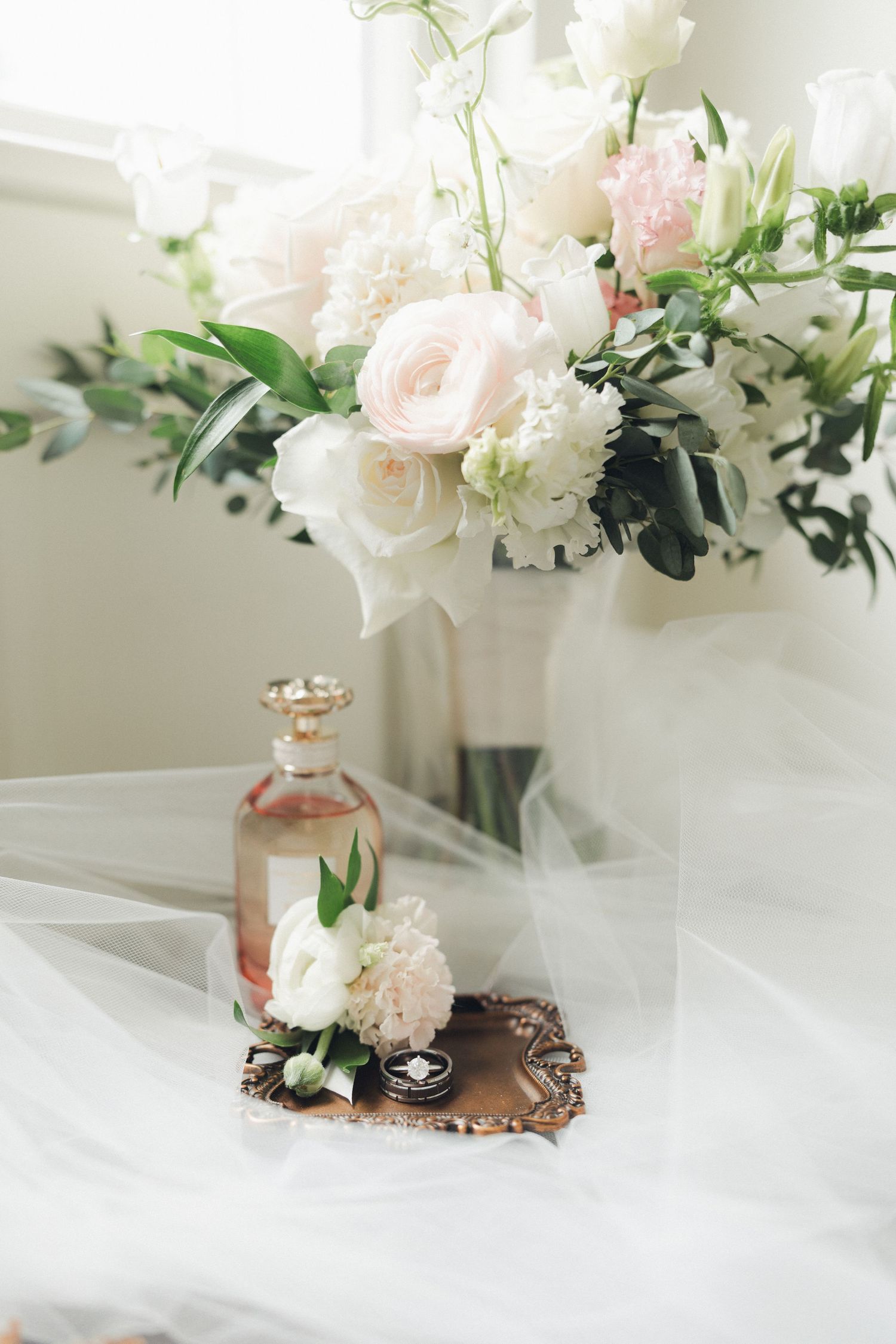 Romantic wedding detail of blush bouquet, vintage perfume bottle, and rings on ornate tray with soft veil.