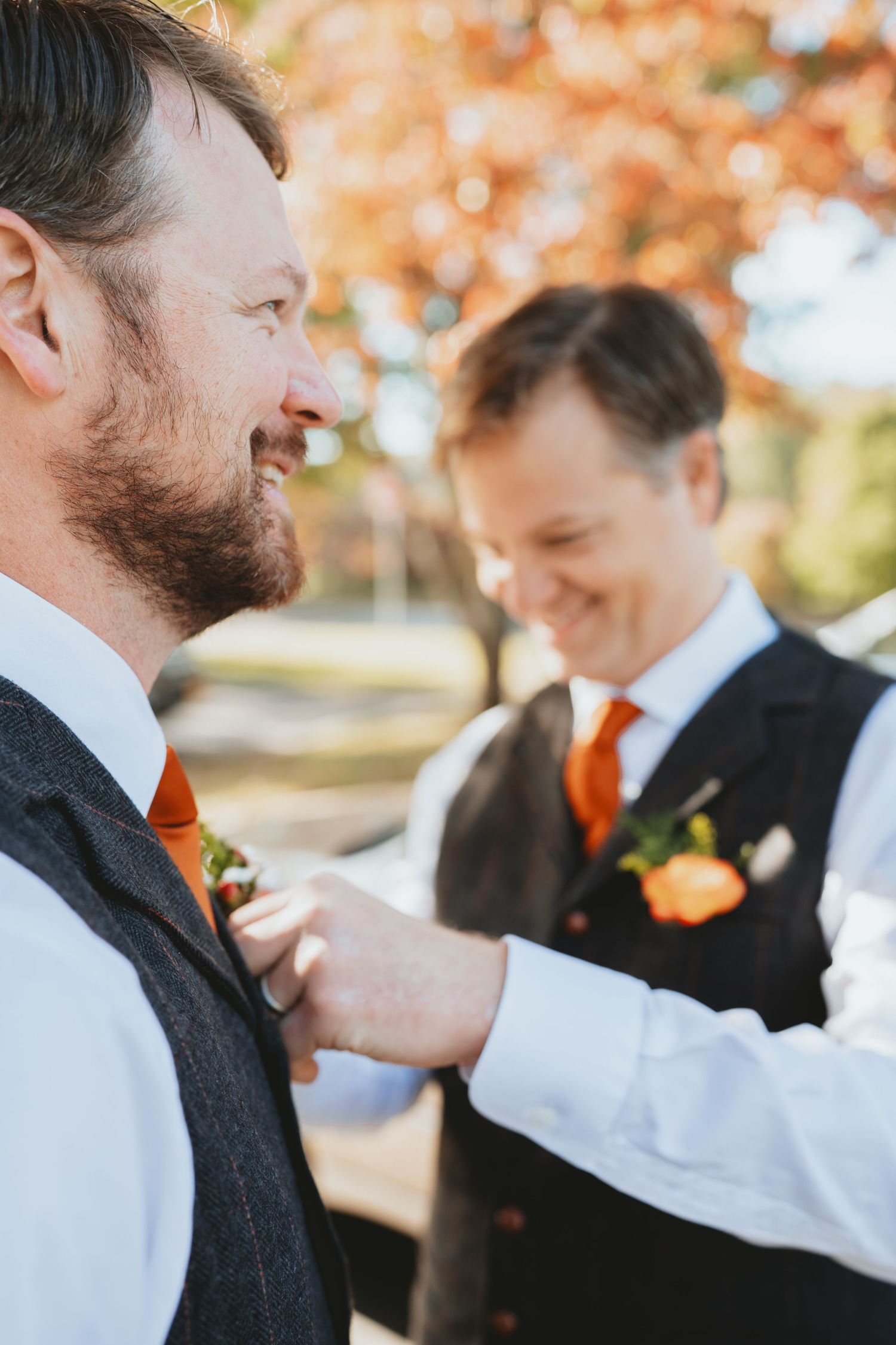 Groomsmen pinning orange boutonniere on groom's grey vest at fall outdoor wedding.