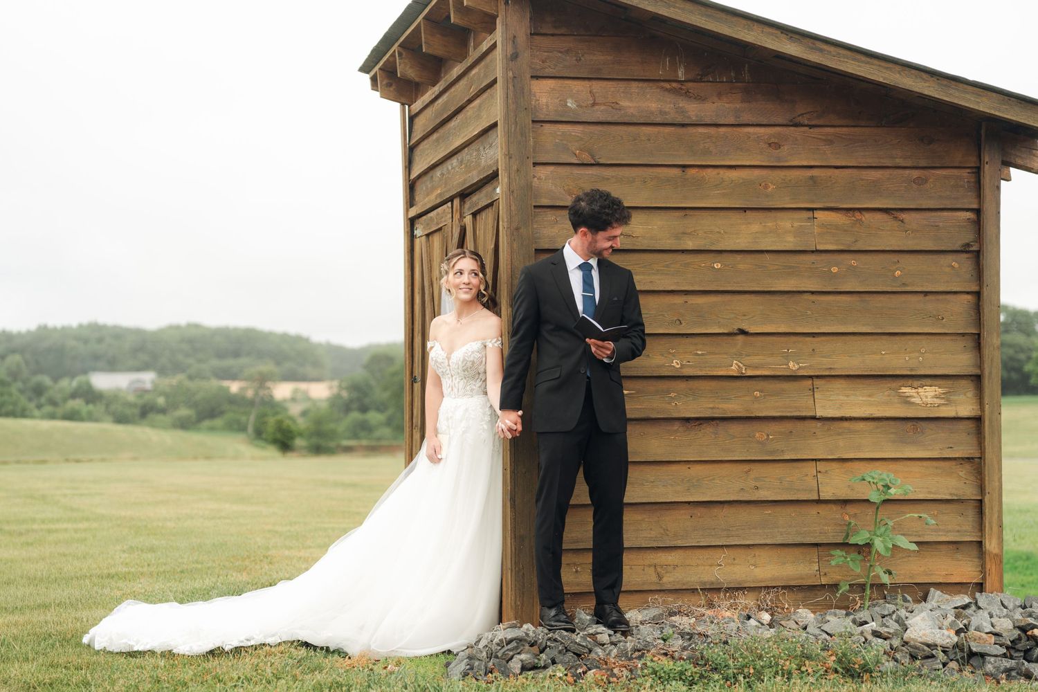 Bride and groom holding hands beside a rustic wooden shed on a green countryside wedding day.