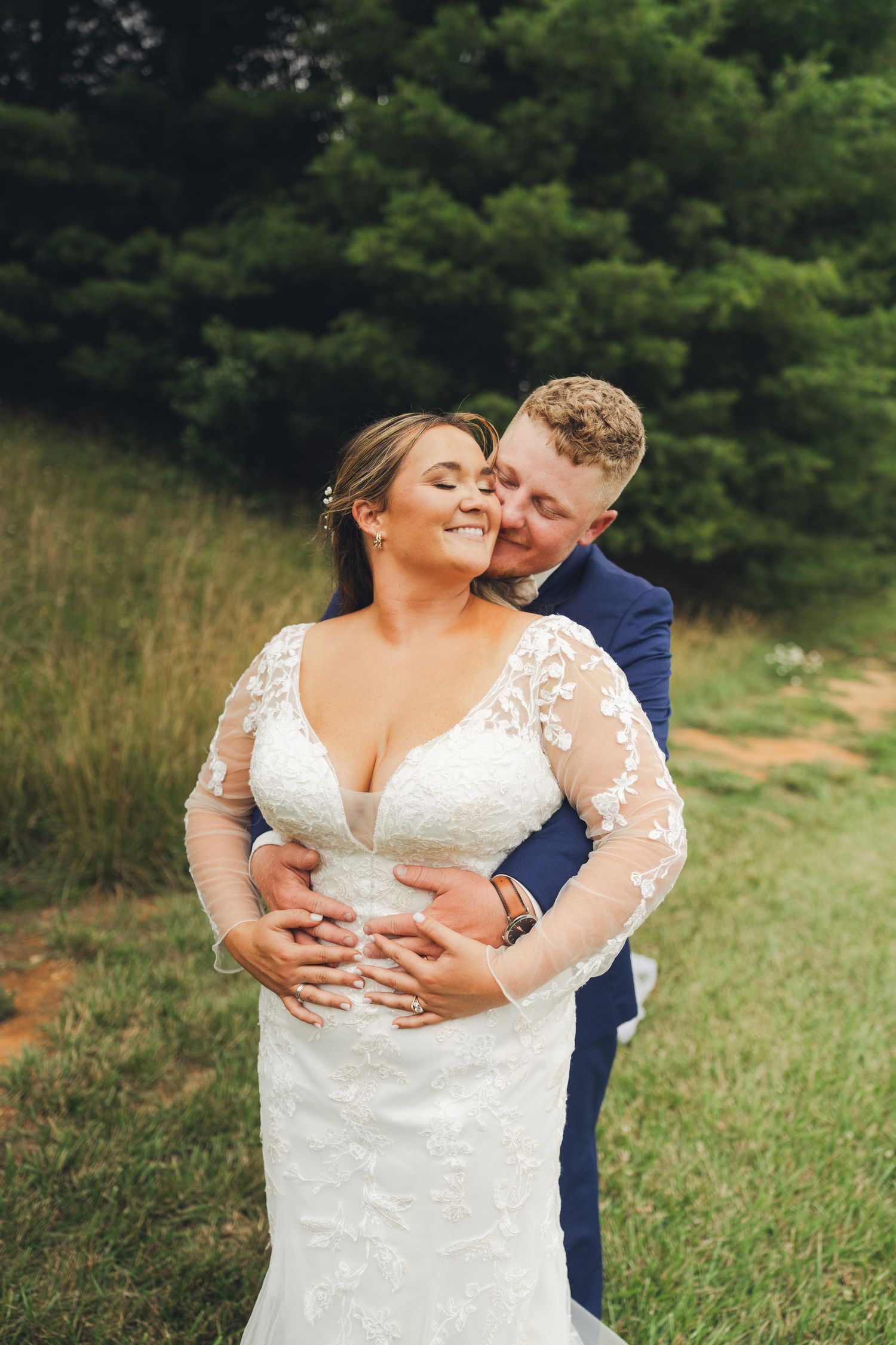 Groom in navy suit embraces bride in lace wedding dress from behind in a lush green outdoor setting.