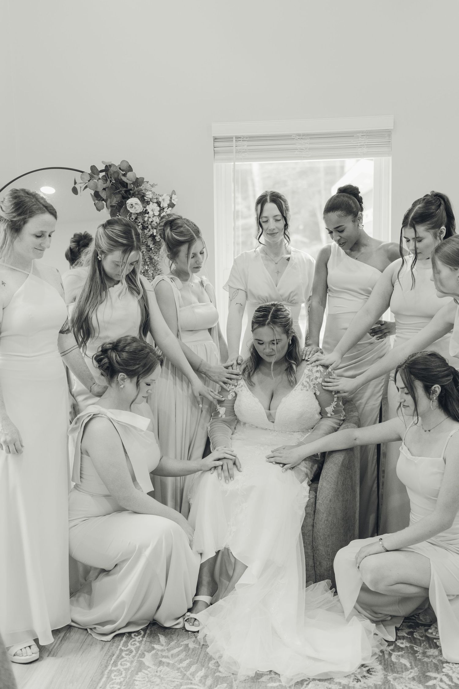 Bridesmaids surrounding and praying over a seated bride in wedding dress, black and white photo.