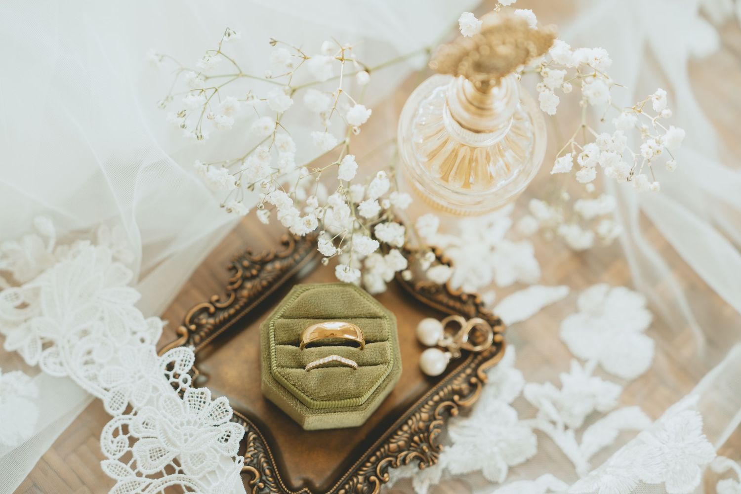 Gold wedding rings in a green velvet box surrounded by baby's breath and lace on a vintage tray.