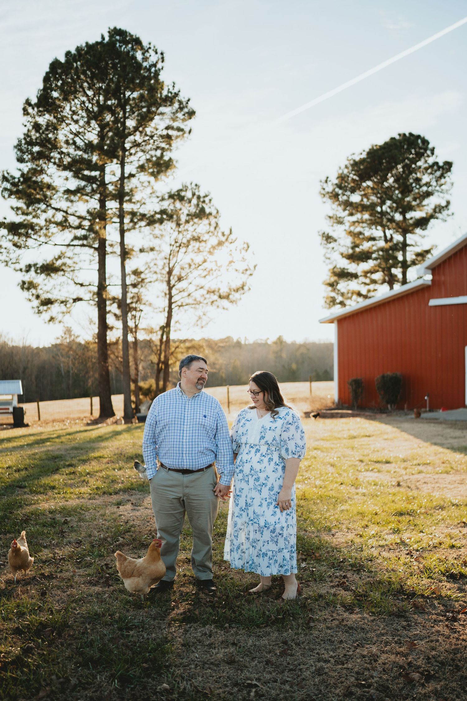 Couple holding hands and smiling at each other near a red barn with chickens at golden hour.