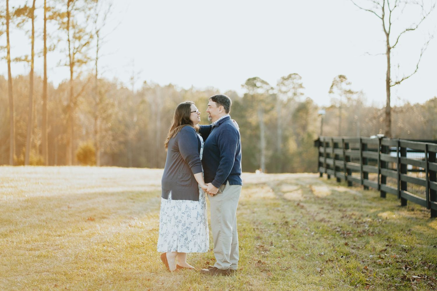 Couple holding hands and facing each other in a sunlit field beside a wooden fence.