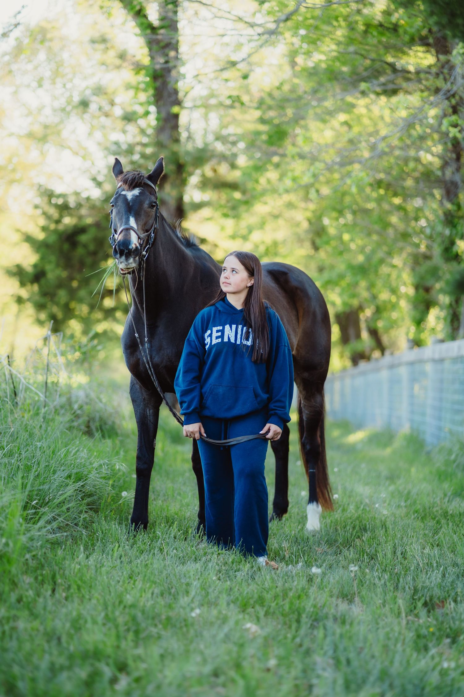 Young woman in blue Senior sweatsuit stands beside a dark horse on a green tree-lined path.