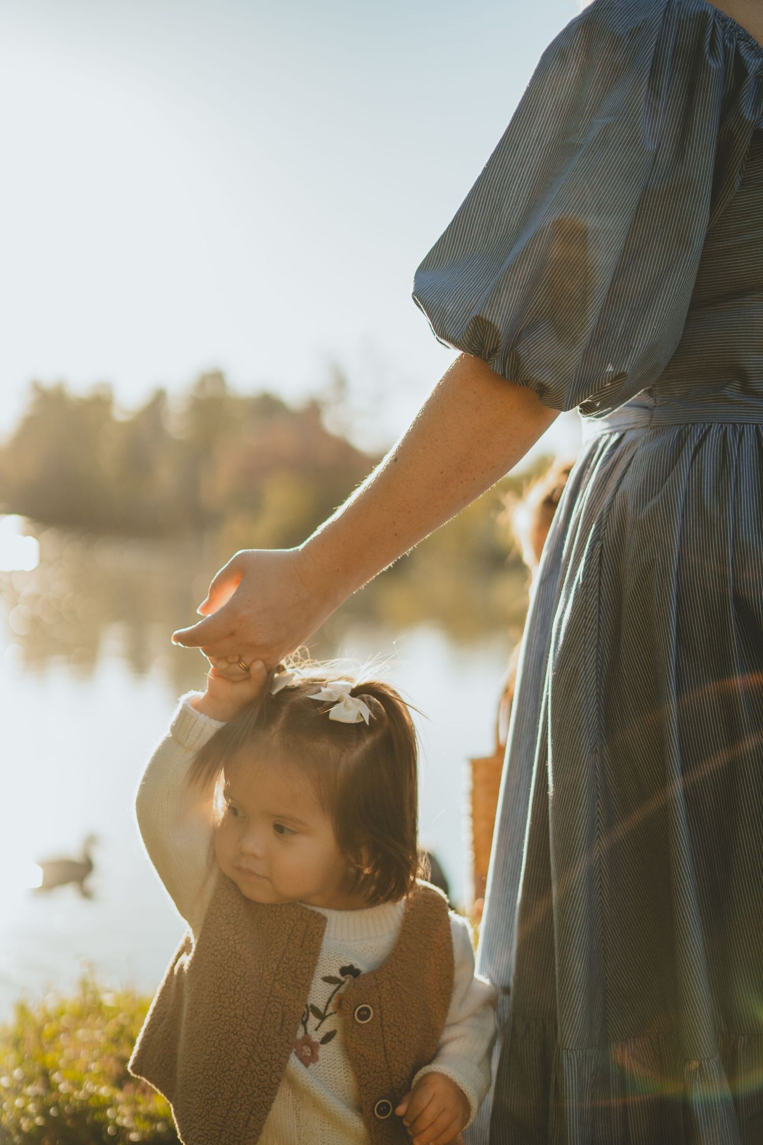 Mother gently holding toddler daughter's hand near sunlit lake with duck in background.