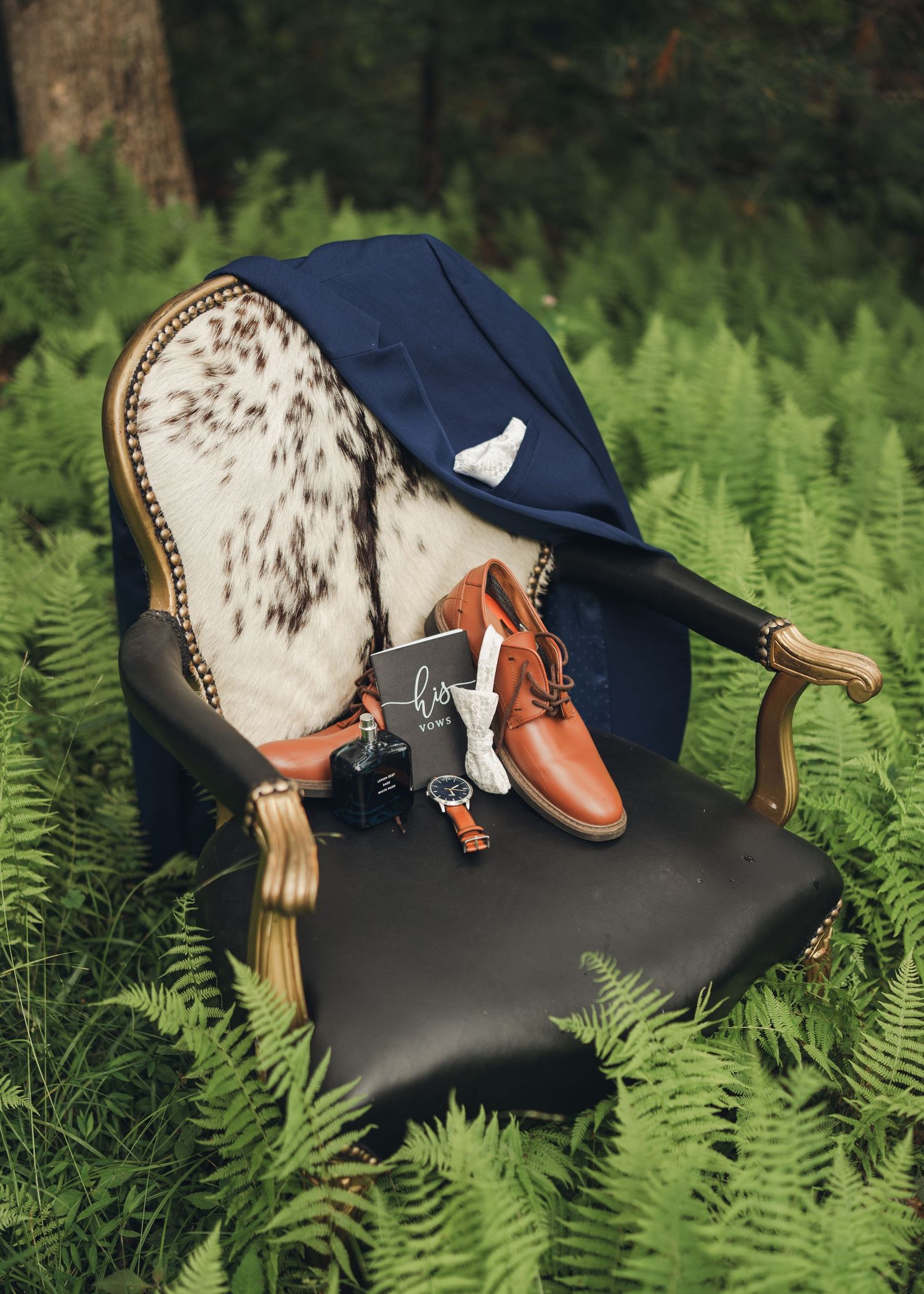 Navy suit jacket and brown leather dress shoes displayed on a vintage chair surrounded by ferns outdoors.