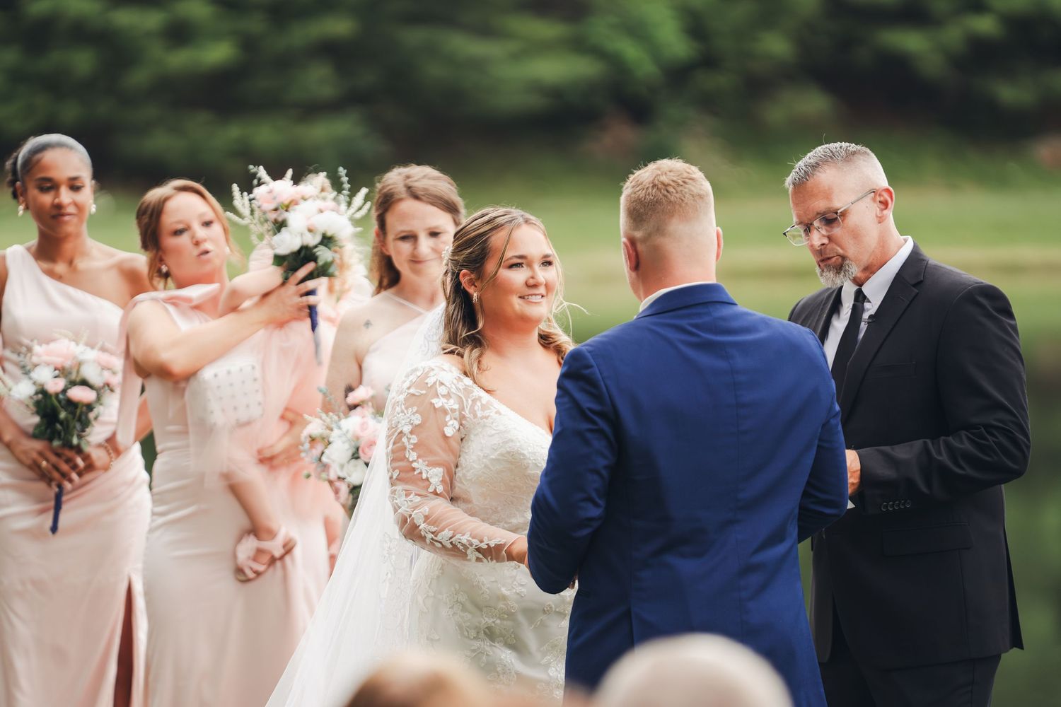 Bride and groom exchange vows outdoors with bridesmaids in blush pink dresses holding bouquets during wedding ceremony.