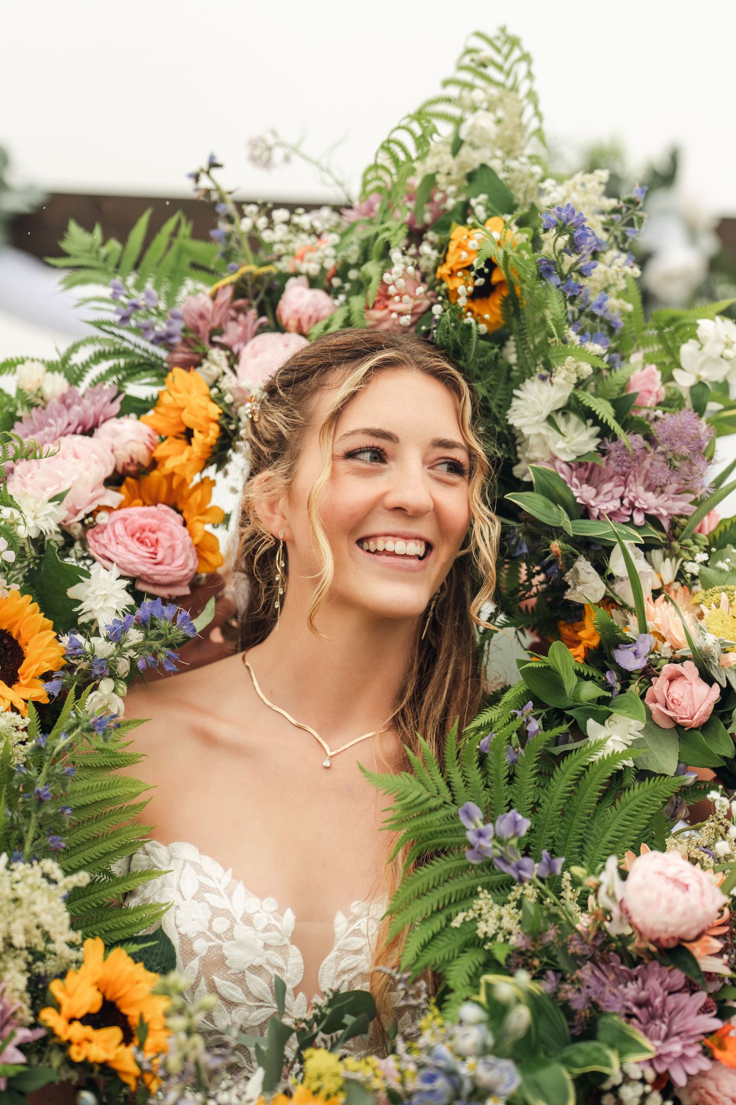 Radiant bride in lace gown smiles joyfully surrounded by a lush colorful floral arrangement with sunflowers and roses.