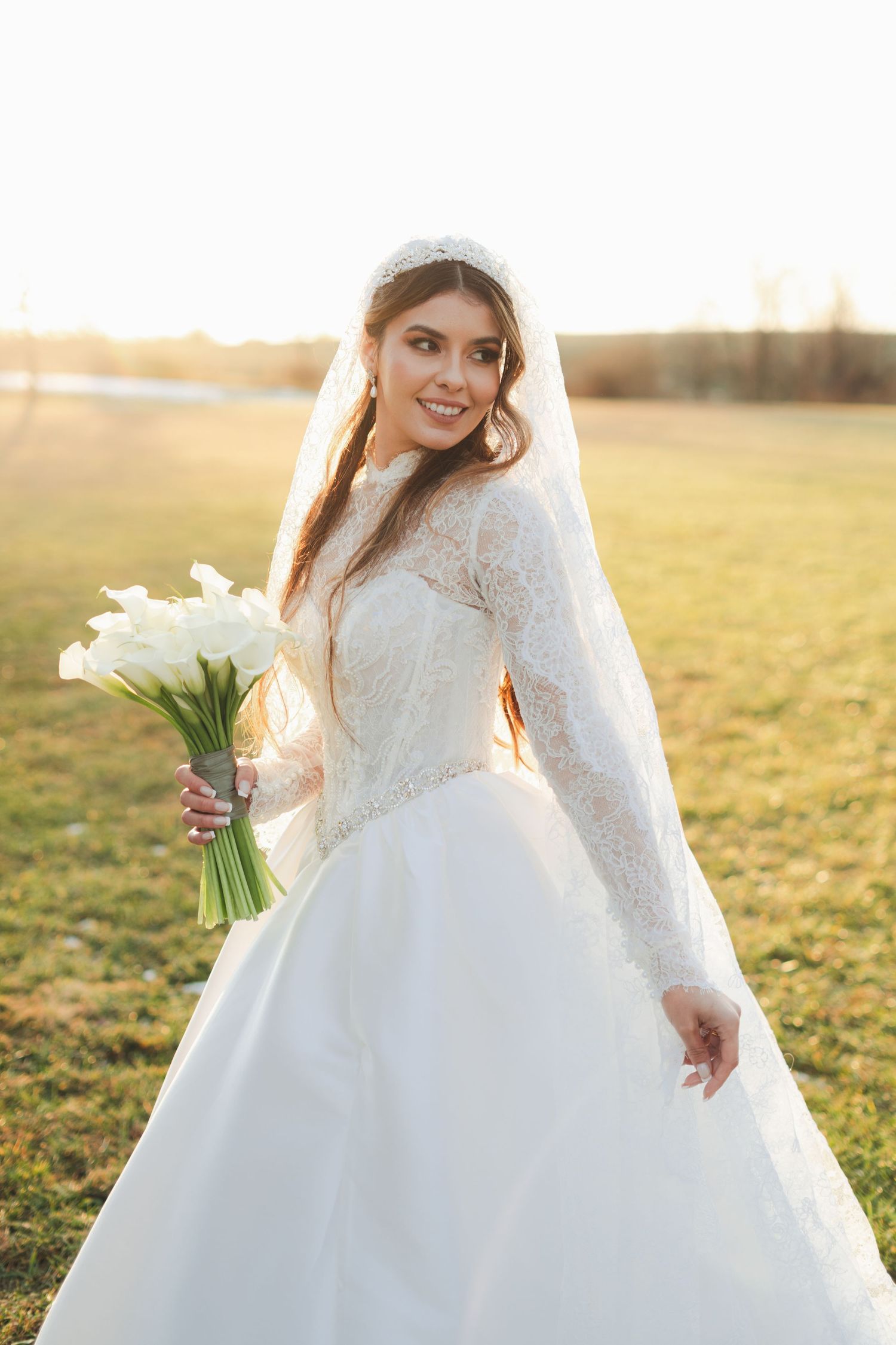 Smiling bride in long-sleeve lace wedding gown and tiara holding calla lily bouquet at sunset.